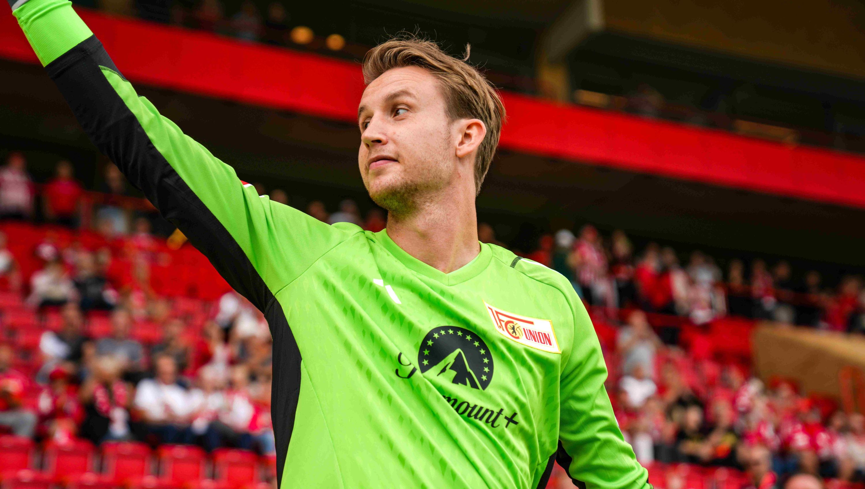 Ein Fußballtorwart in grüner Trikot steht lächelnd auf dem Spielfeld und winkt den Fans zu.