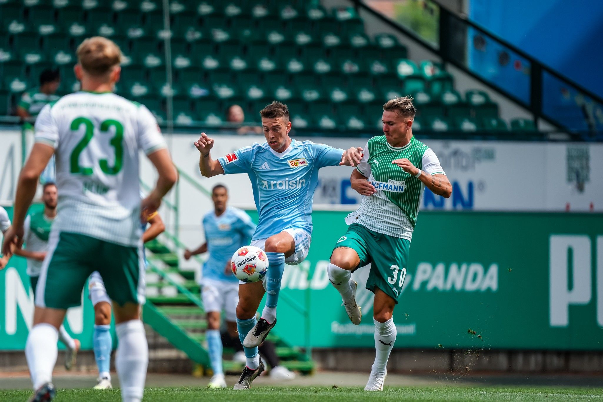 Zwei Fußballspieler im direkten Duell auf dem Spielfeld, einer im blauen Trikot und der andere im grünen Trikot.