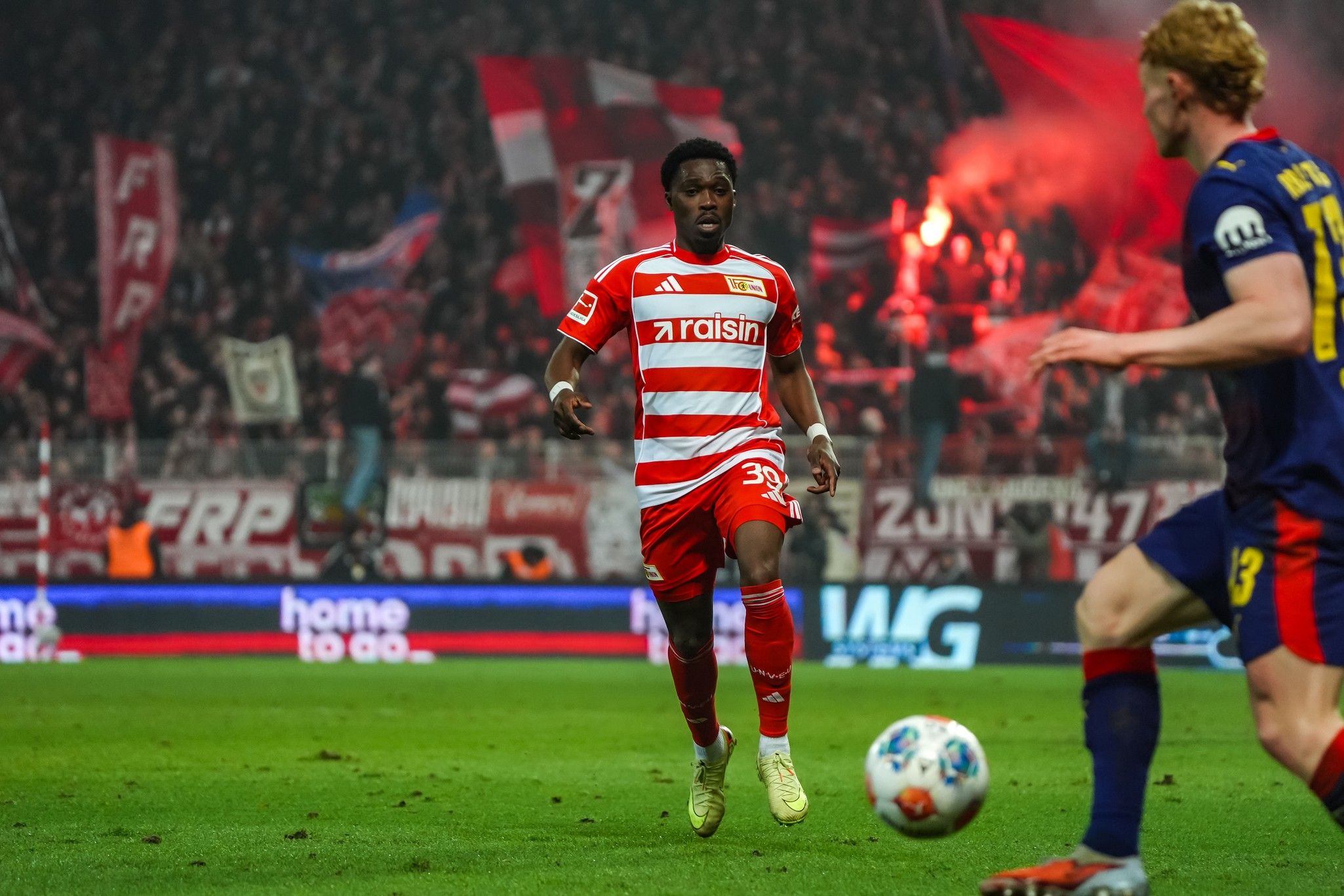 Ein Fußballspieler in rot-weißer Trikotausrüstung dribbelt den Ball, während die Menge im Hintergrund jubelt.