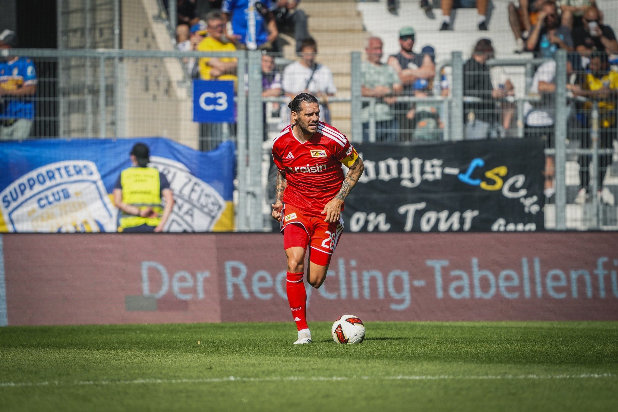 Ein Fußballspieler in rotem Trikot läuft mit dem Ball auf einem Stadionfeld, Zuschauer im Hintergrund.