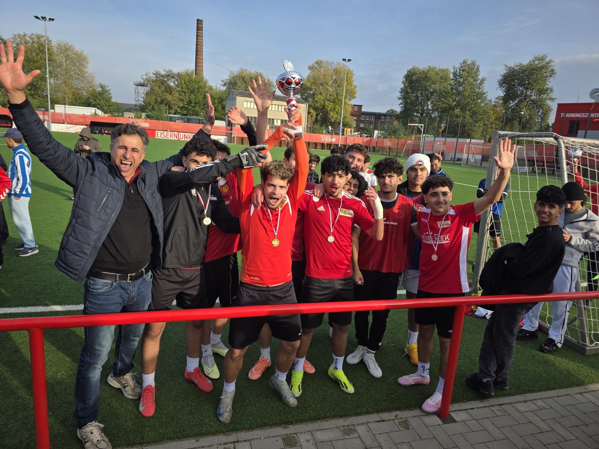 Gruppe junger Fußballspieler in roten Trikots feiert mit jubelnden Gesten und Medaillen auf einem Sportplatz.