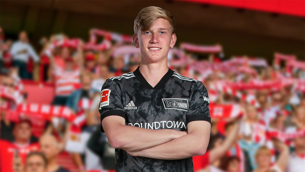 Ein junger Fußballspieler in einem schwarzen Trikot mit verschränkten Armen, lächelt vor einer jubelnden Menschenmenge.