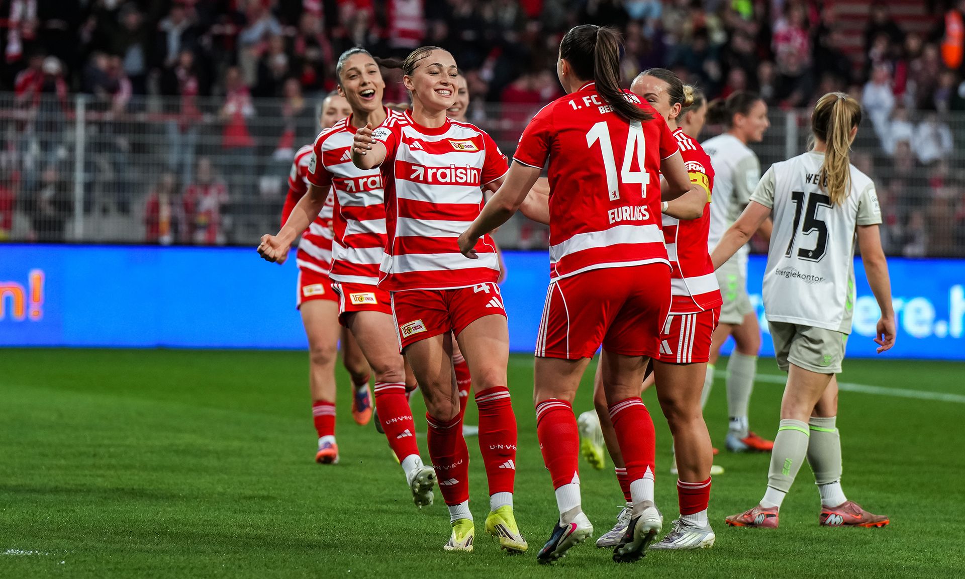 Spielerinnen des Fußballteams in rot-weißen Trikots feiern einen Torerfolg auf dem Spielfeld.