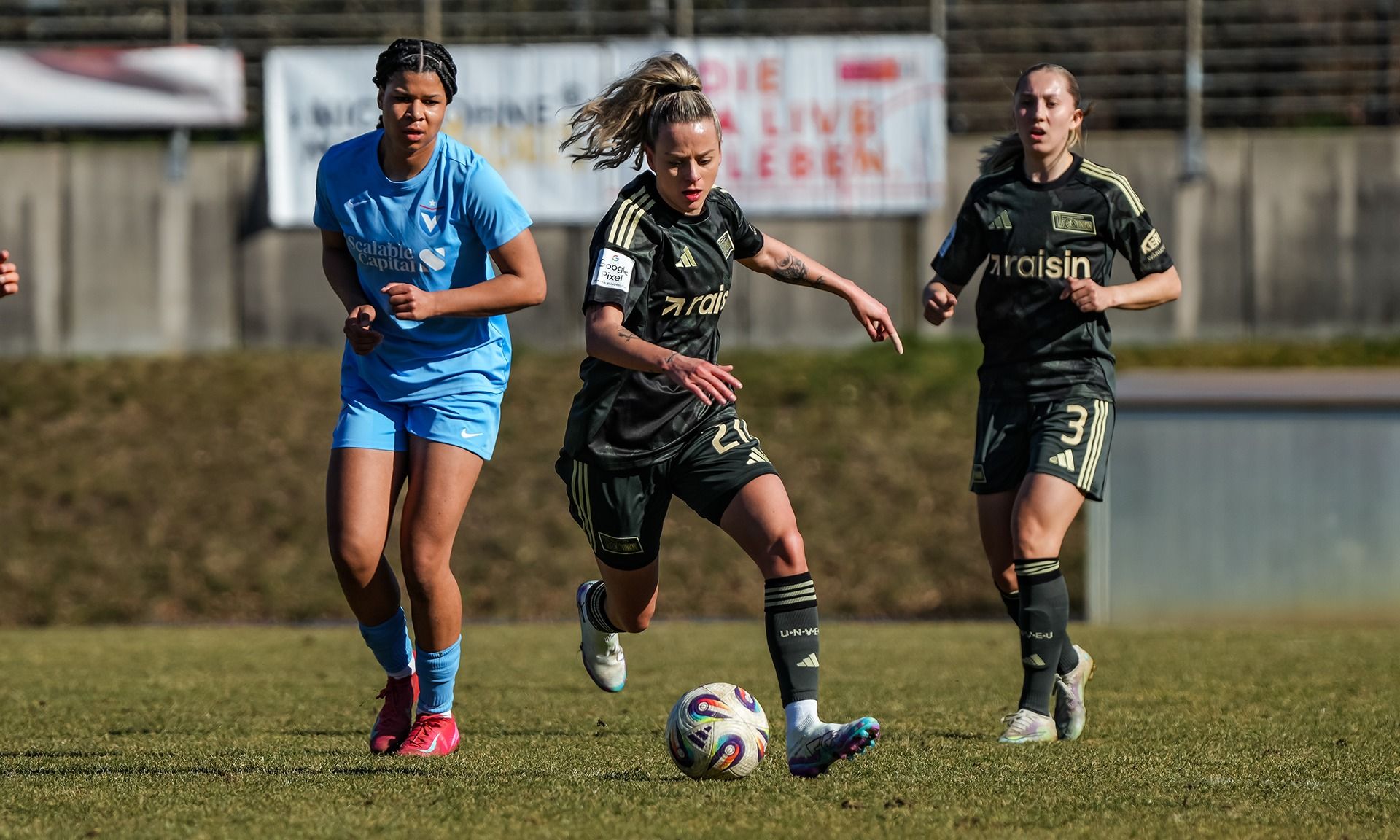 Eine Fußballspielerin in schwarzer Uniform dribbelt den Ball, während zwei Gegnerinnen in blauen Trikots sie verfolgen.