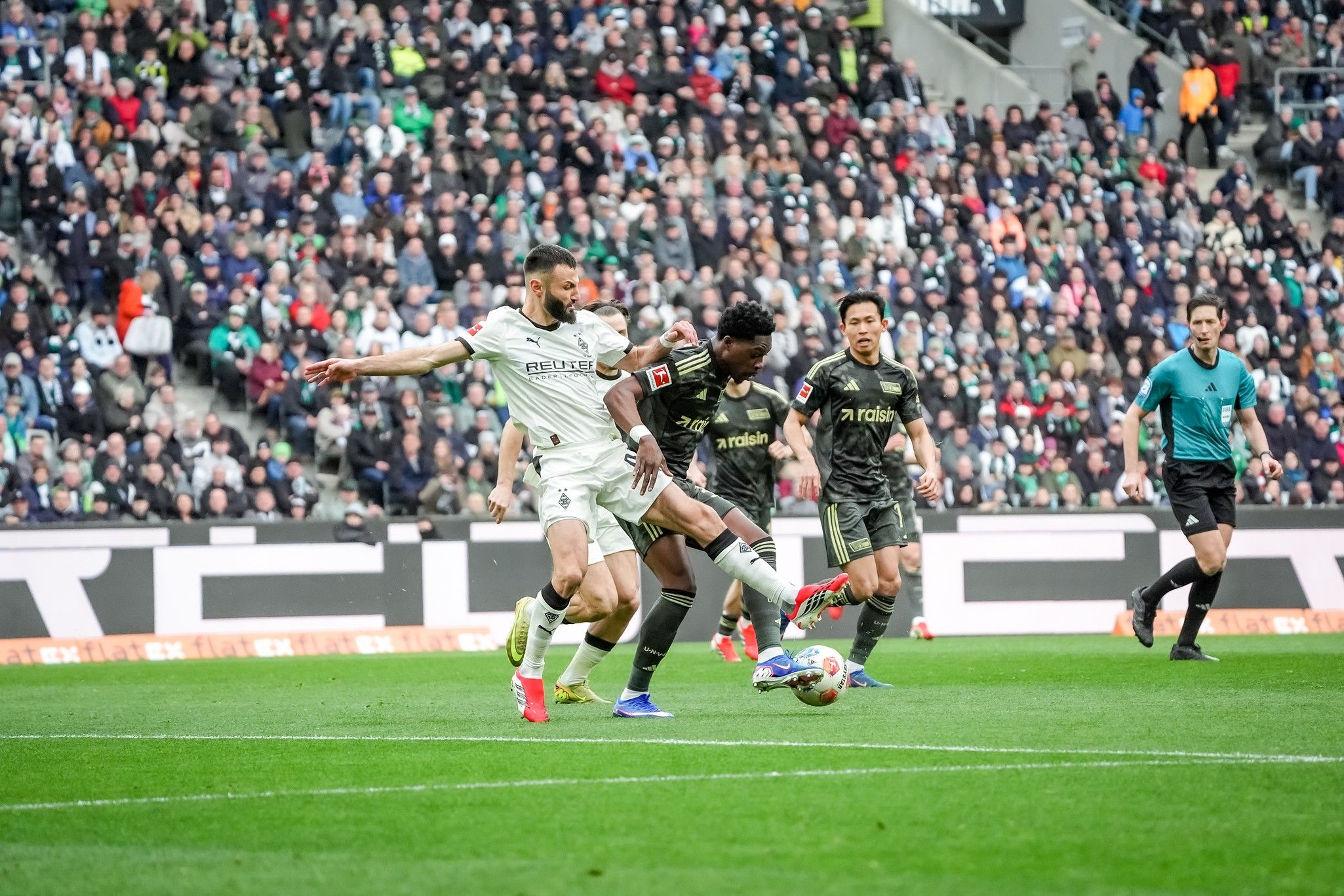 Spielszene im Fußball mit Spielern in Sportswear, die um den Ball kämpfen, während Zuschauer im Hintergrund anfeuern.