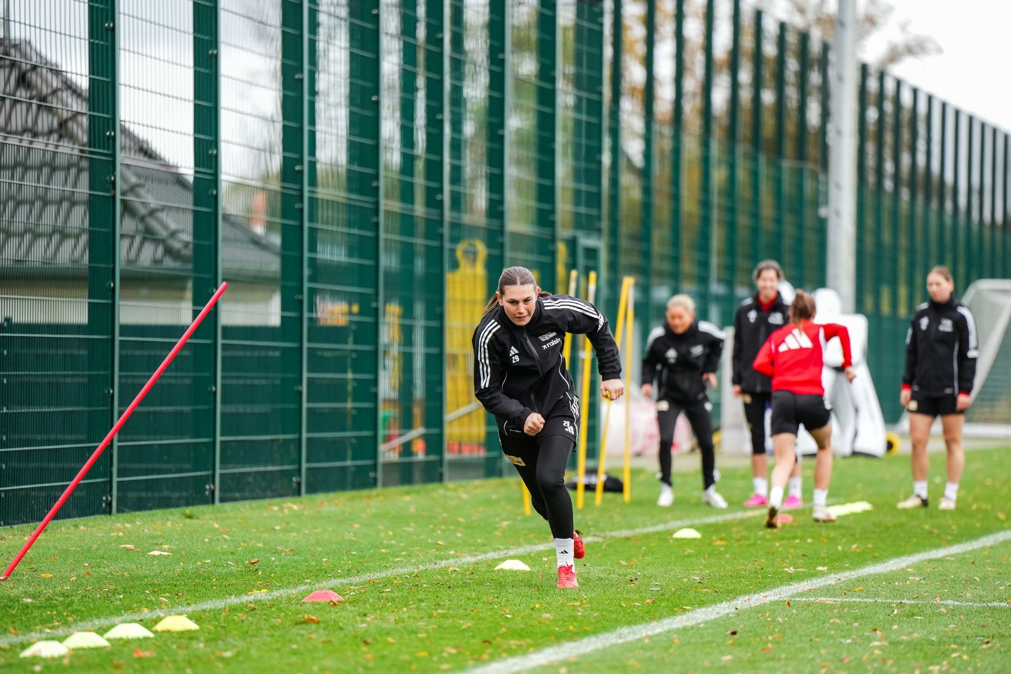 Eine Sportlerin sprintet auf einem grünen Trainingsplatz, während andere Spielerinnen im Hintergrund Übungen machen.