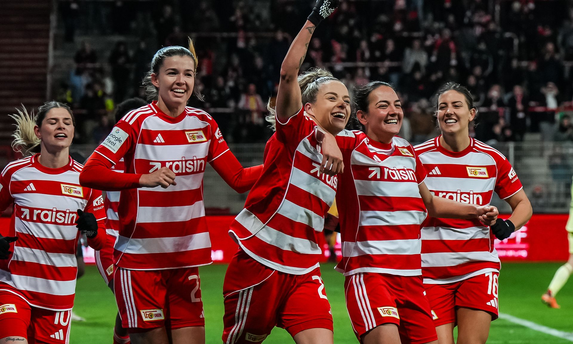 Frauenfußballmannschaft mit rot-gestreiften Trikots feiert einen Torerfolg im Stadion. Spielerinnen sind fröhlich und jubeln.