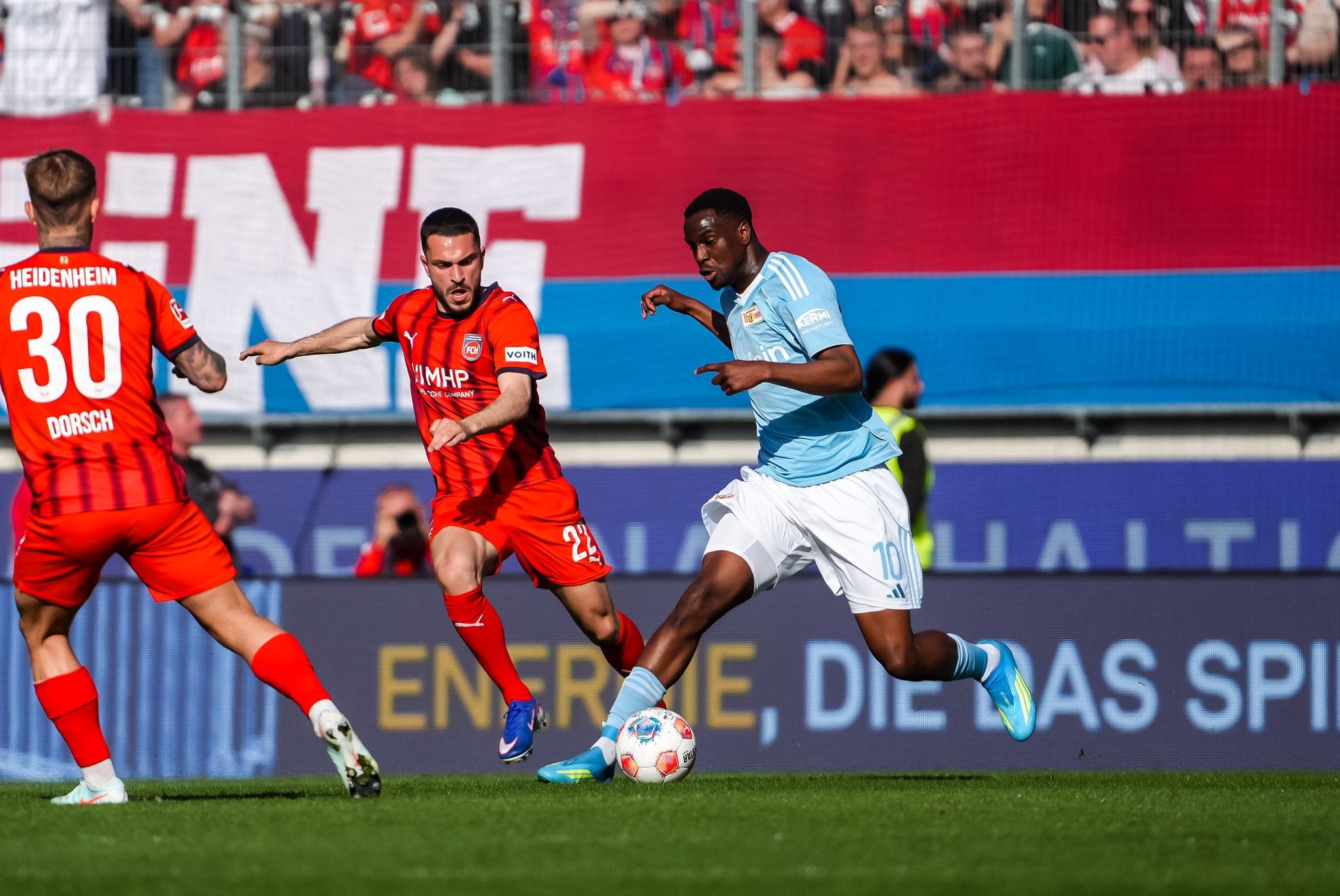 Ein Fußballspieler in blauer Trikotfarbe dribbelt den Ball, während zwei Spieler in roten Trikots ihn verfolgen.
