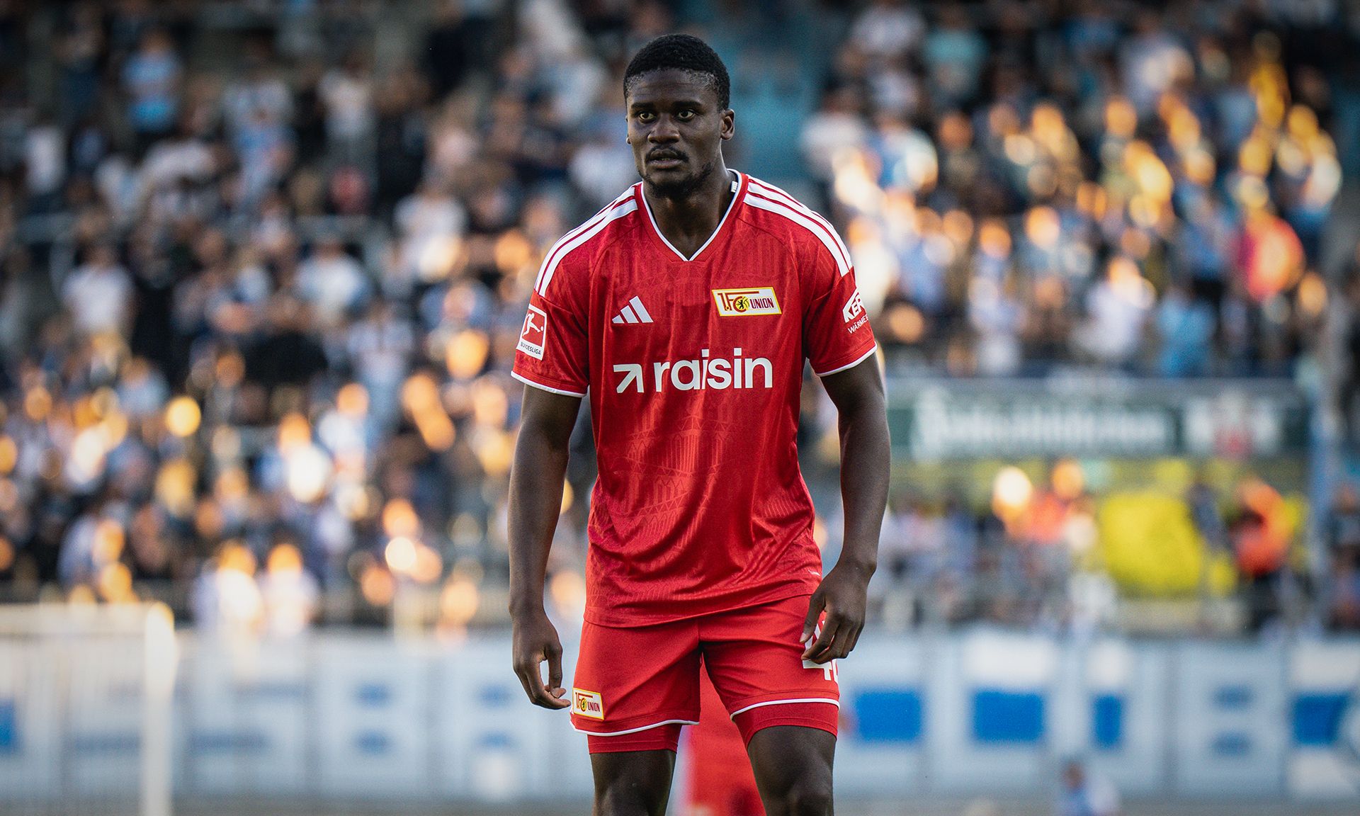 Spieler in roter Fußballuniform steht auf dem Spielfeld, im Hintergrund ist eine Menschenmenge zu sehen.
