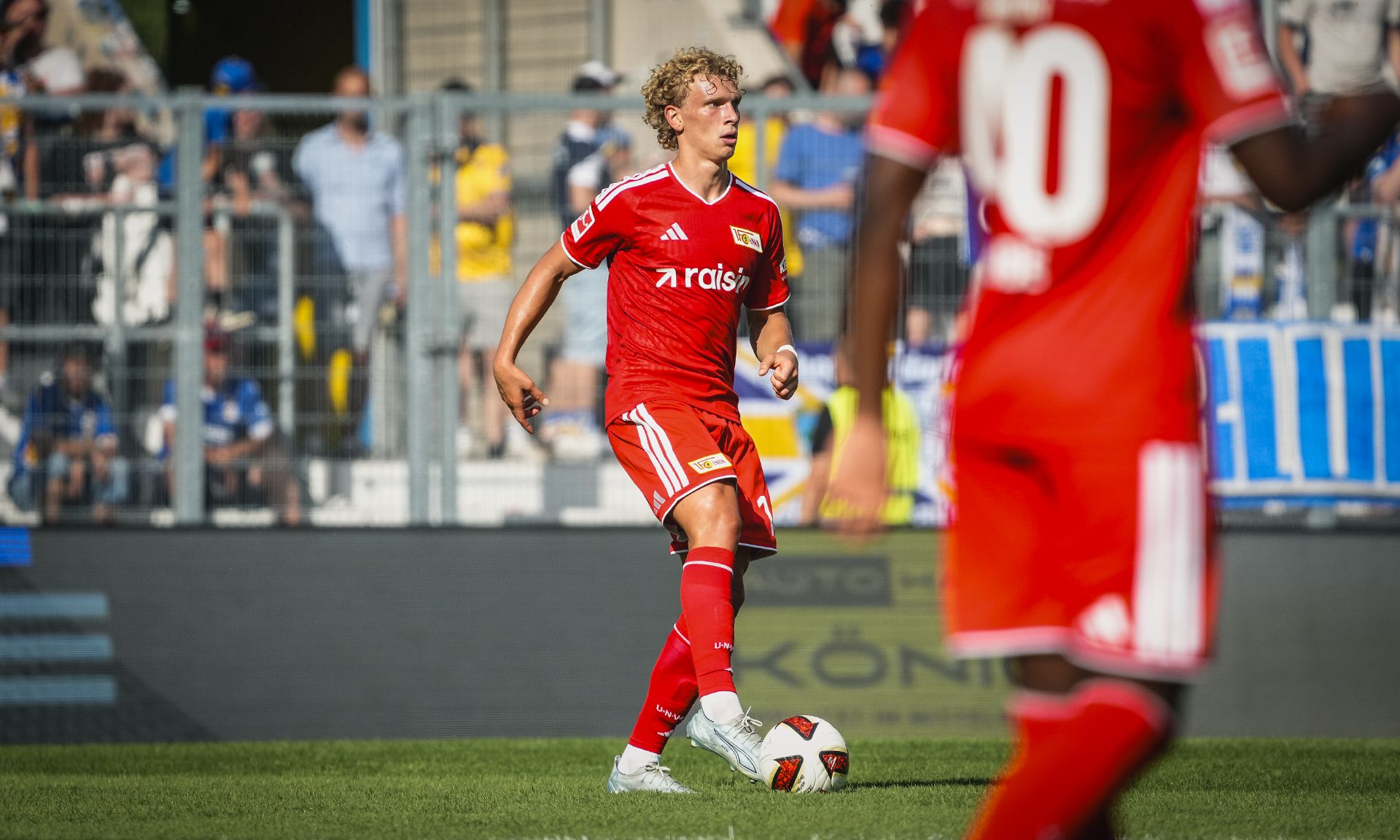 Ein Fußballspieler in roter Ausrüstung steht auf dem Spielfeld und hat den Ball am Fuß, im Hintergrund Zuschauer.
