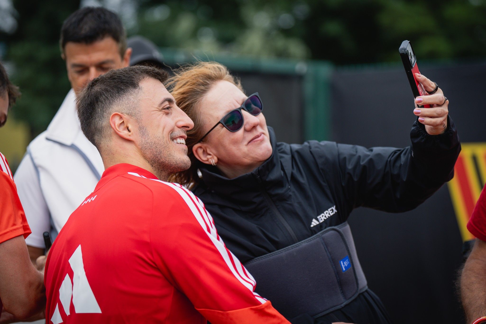 Zwei Personen machen ein Selfie, während sie lächeln. Eine Person trägt ein rotes Sporttrikot, die andere eine schwarze Jacke.