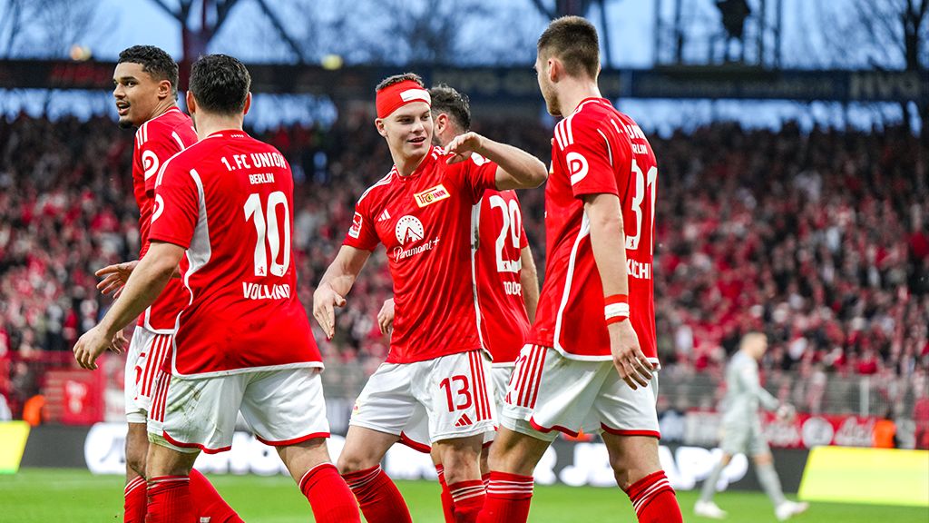 Players of 1. FC Union Berlin celebrate a goal in front of cheering fans in a stadium. They all wear red jerseys.