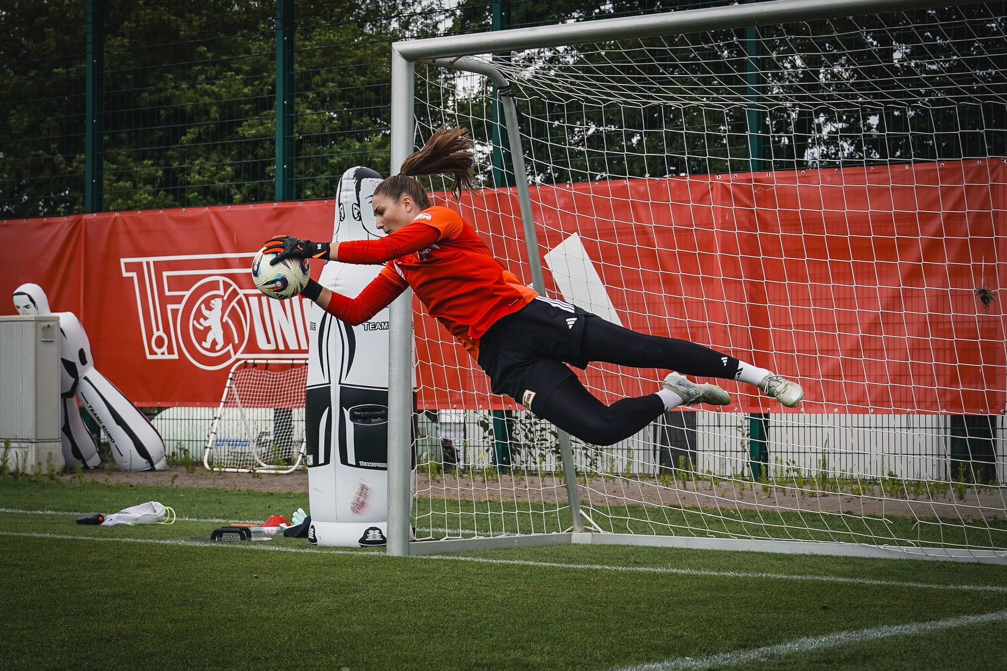 Torhüterin in orangefarbener Ausrüstung springt, um einen Ball im Fußballtor zu halten. Hintergrund mit Vereinsbanner und Torpfosten.