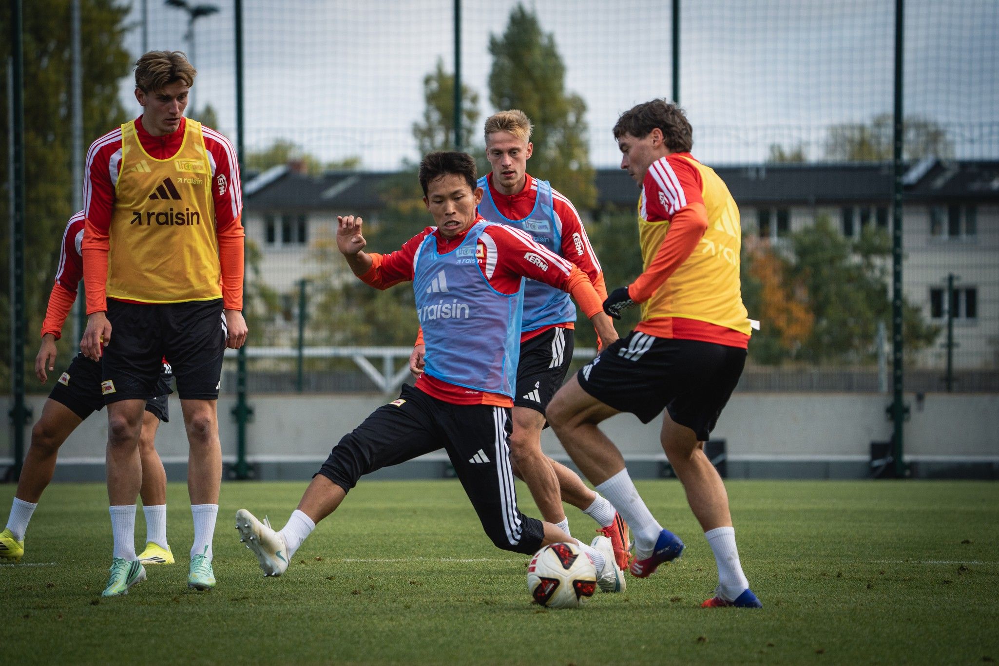 Vier Fußballspieler trainieren auf einem Platz, einer führt einen Torschuss aus, während die anderen beobachten.