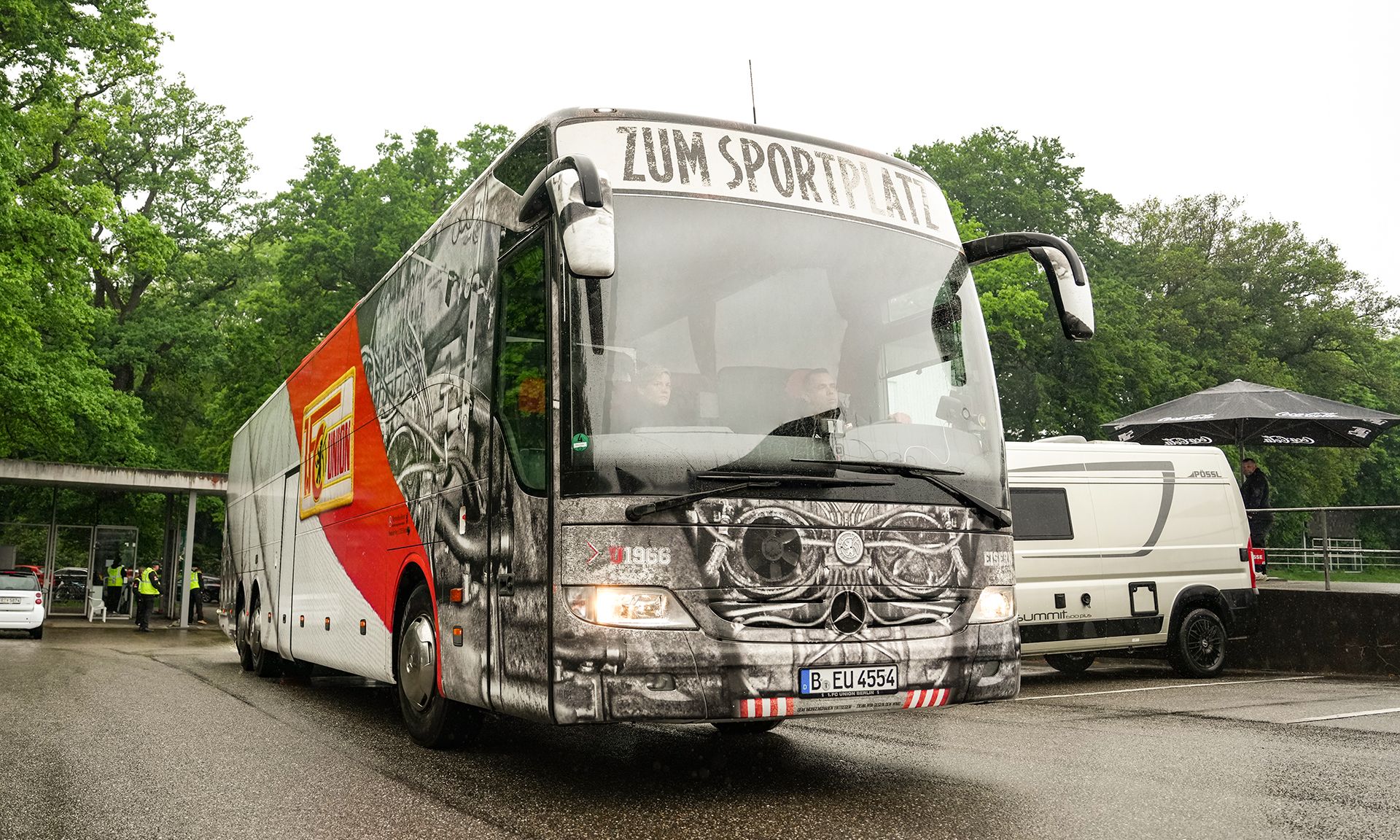 Ein Bus mit auffälliger Grafik fährt auf einer regnerischen Straße, umgeben von grünen Bäumen und parkenden Fahrzeugen.