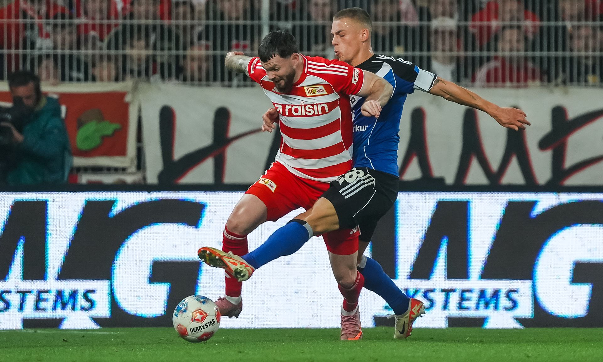 Zwei Fußballspieler im Duell, einer fällt zu Boden, während der andere ihn herausfordert. Stadionatmosphäre im Hintergrund.