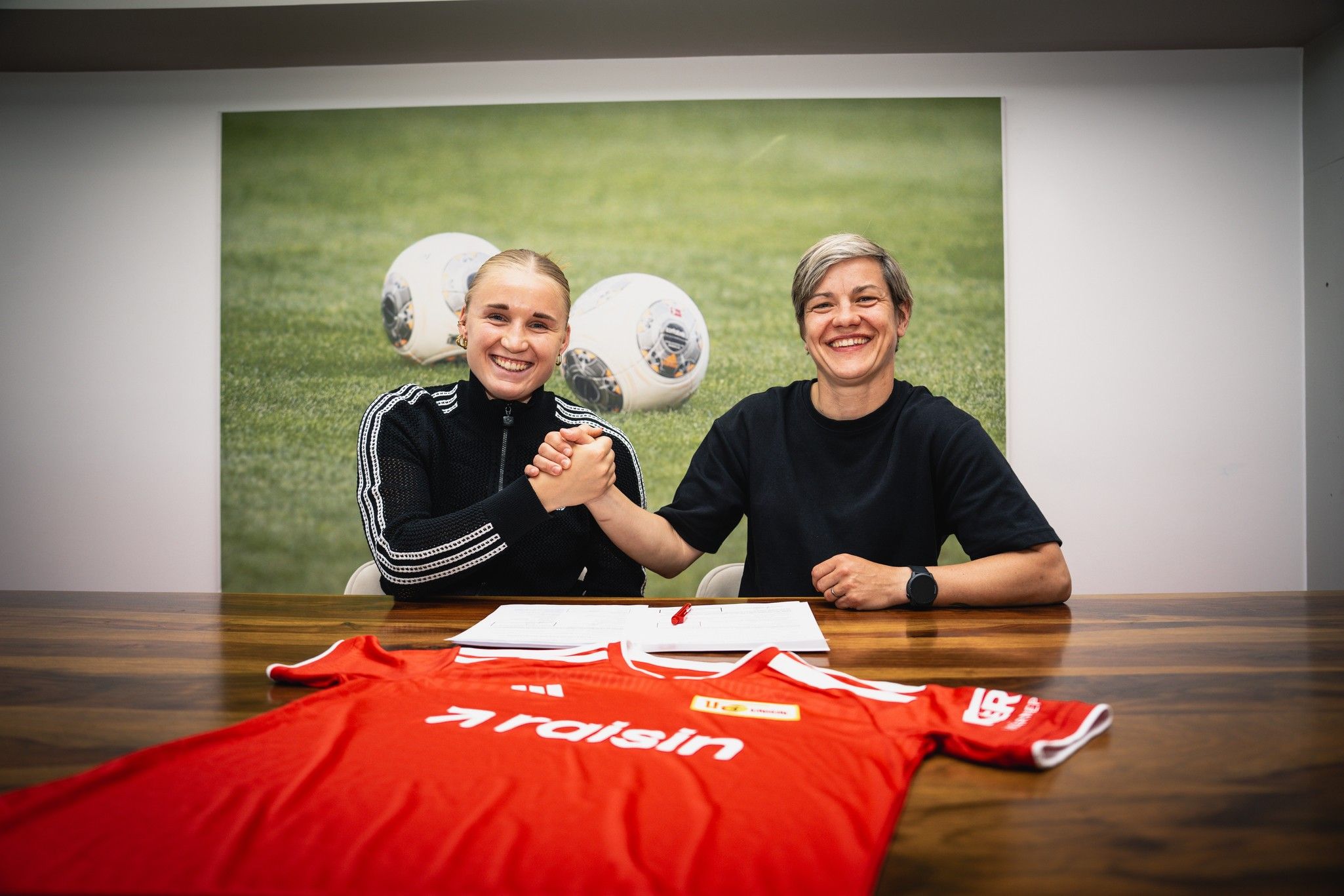 Zwei Frauen schütteln sich die Hand über einem Tisch, auf dem ein rotes Trikot liegt, während ein Fußballfeld im Hintergrund sichtbar ist.