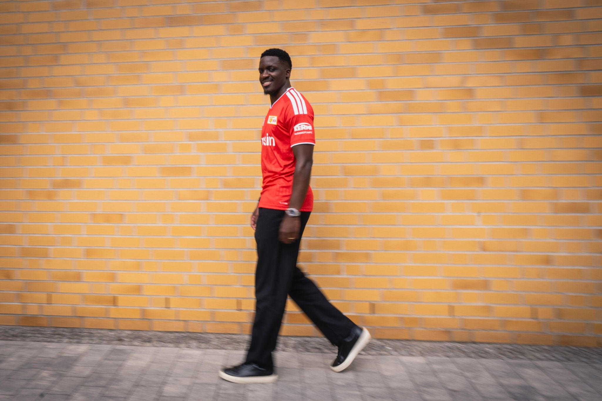 Ein Mann in einem roten Sporttrikot geht lächelnd an einer gelben Backsteinmauer entlang.