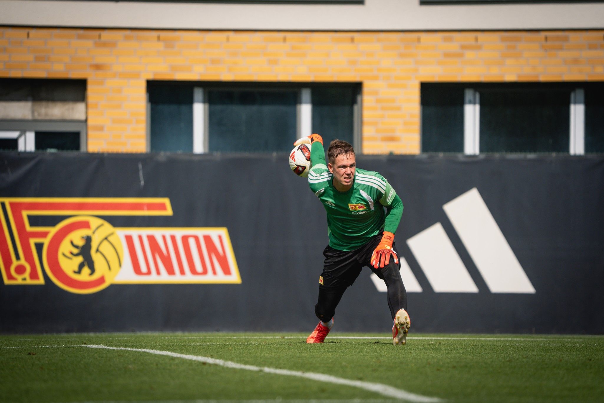Torwart in grünem Trikot bereitet sich auf einen Wurf vor, mit einem Fußball in der Hand, im Training auf einem Sportplatz.