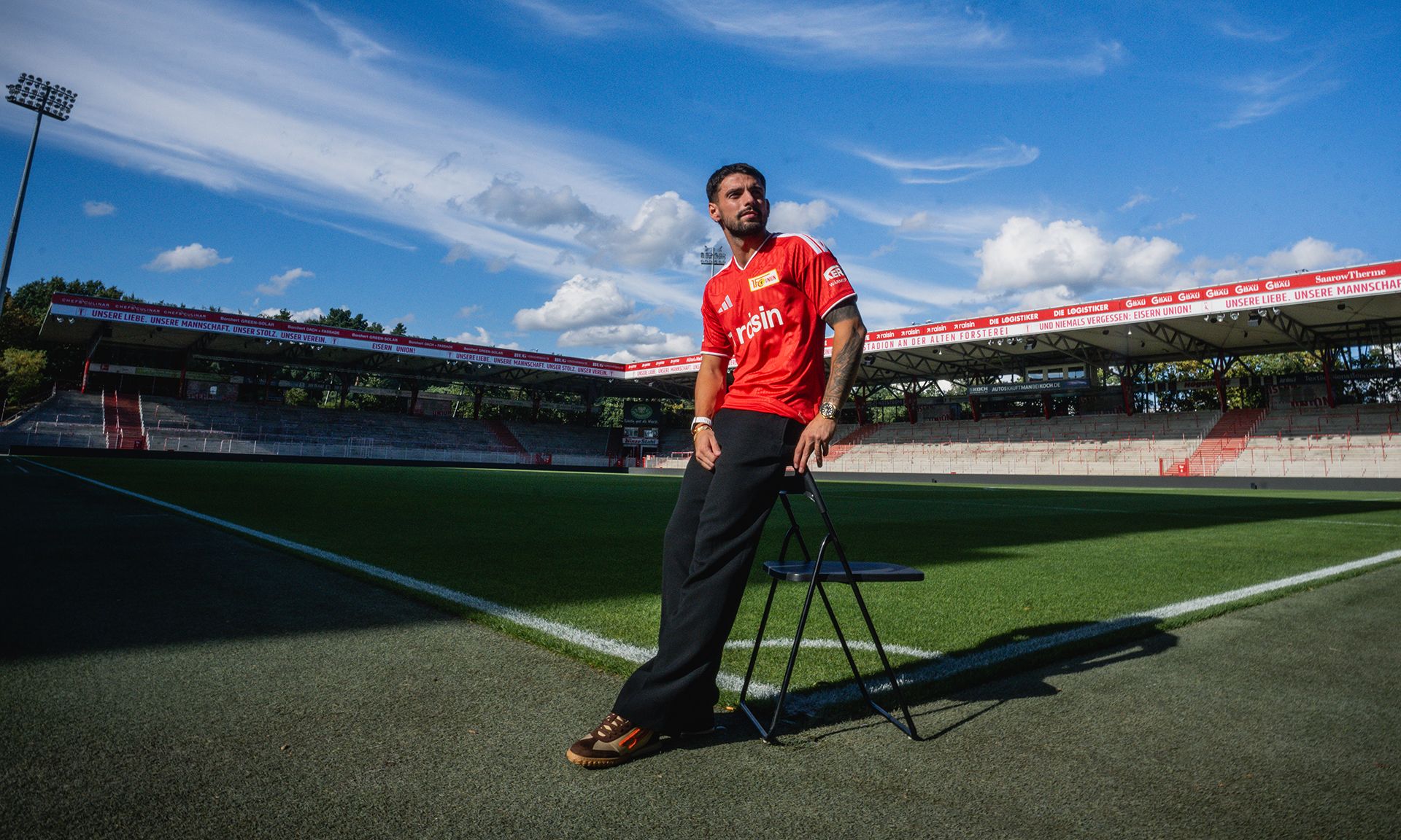 Ein Fußballspieler in rotem Trikot sitzt auf einem Stuhl im Stadion, umgeben von Rasen und blauen Himmel.