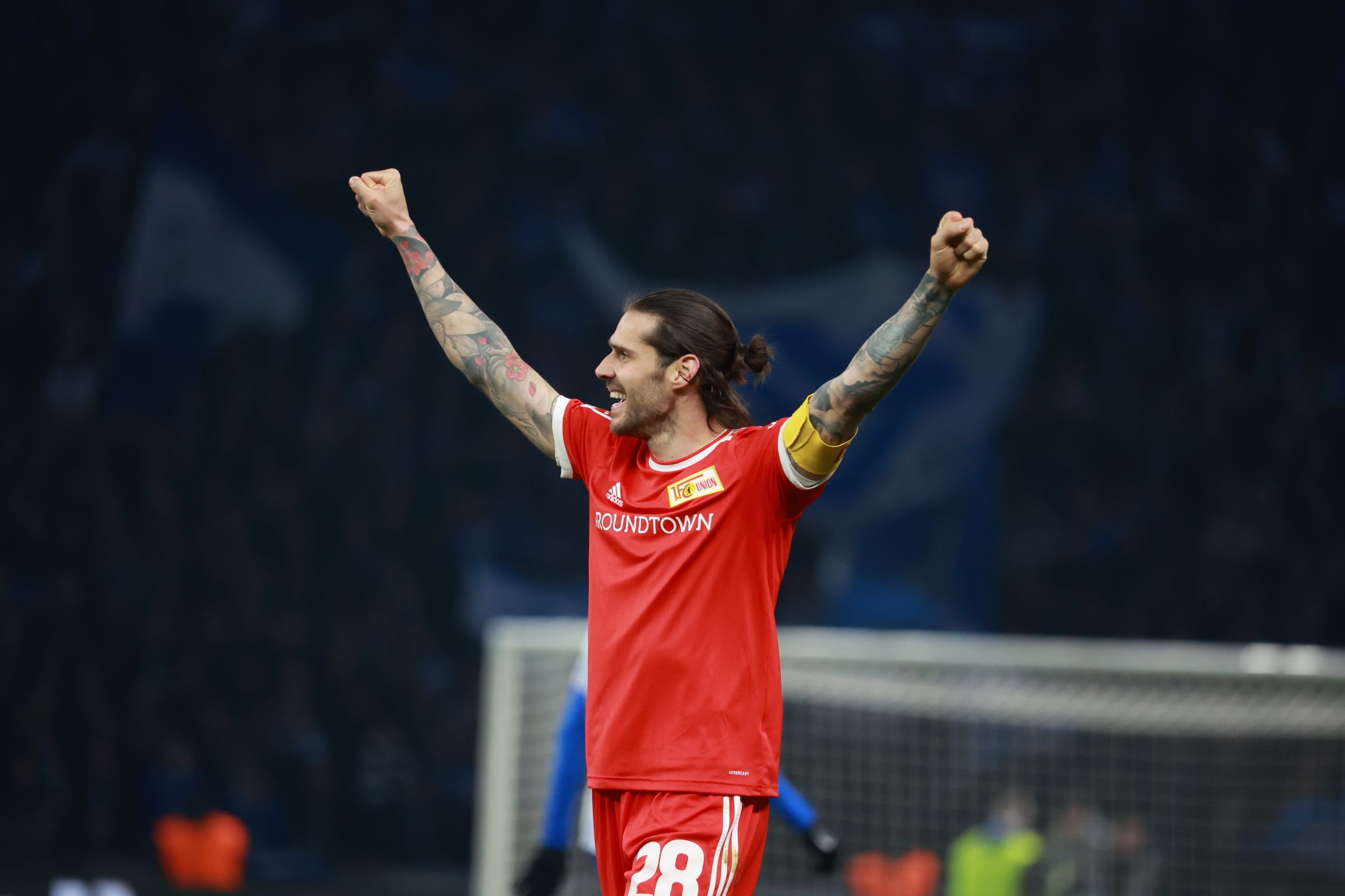Ein Fußballspieler in roter Uniform mit erhobenen Armen jubelt vor einer jubelnden Menge im Stadion.