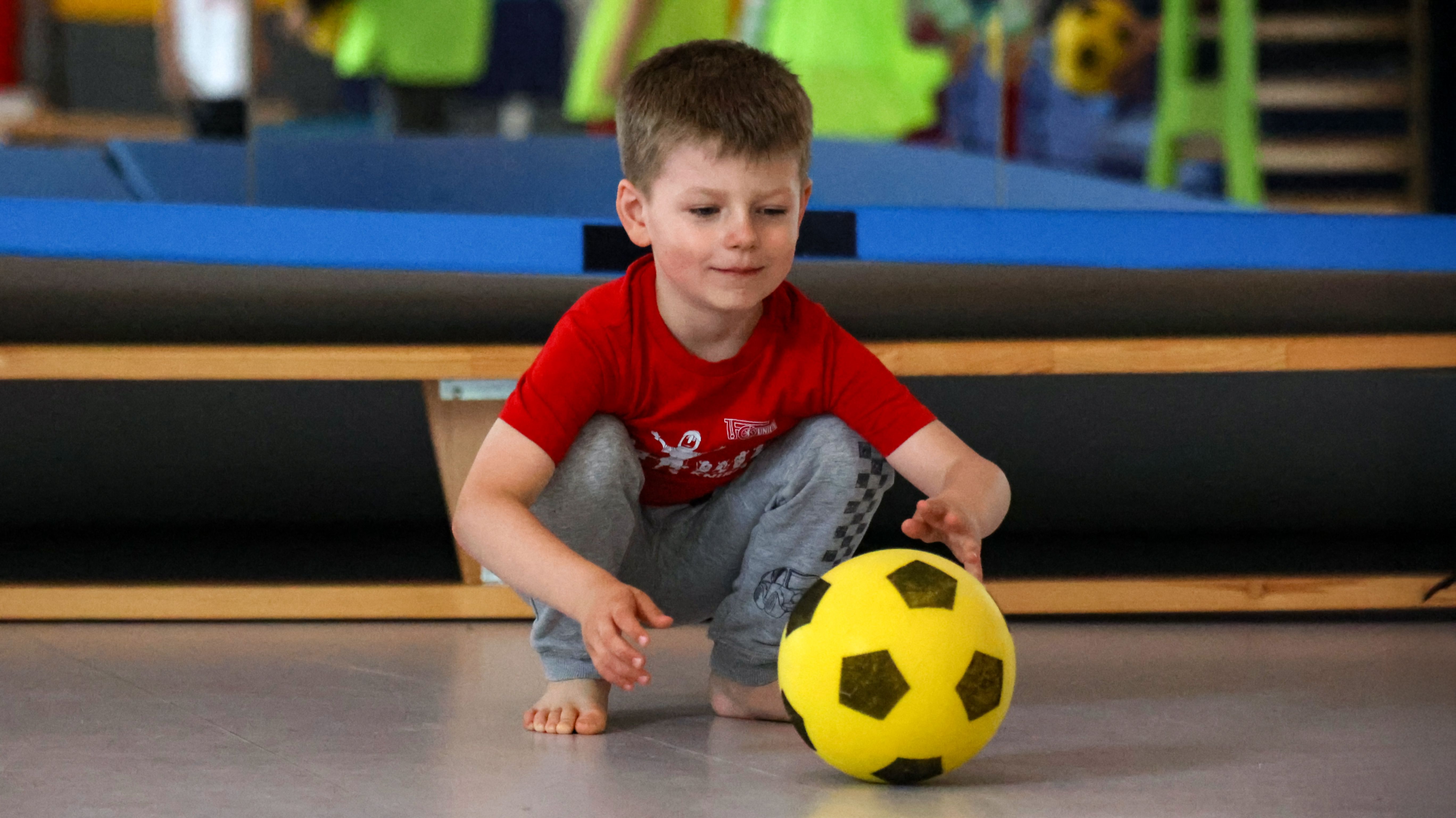 Ein Junge in rotem Shirt und grauen Hosen spielt auf einem Sportboden mit einem gelben Fußball.