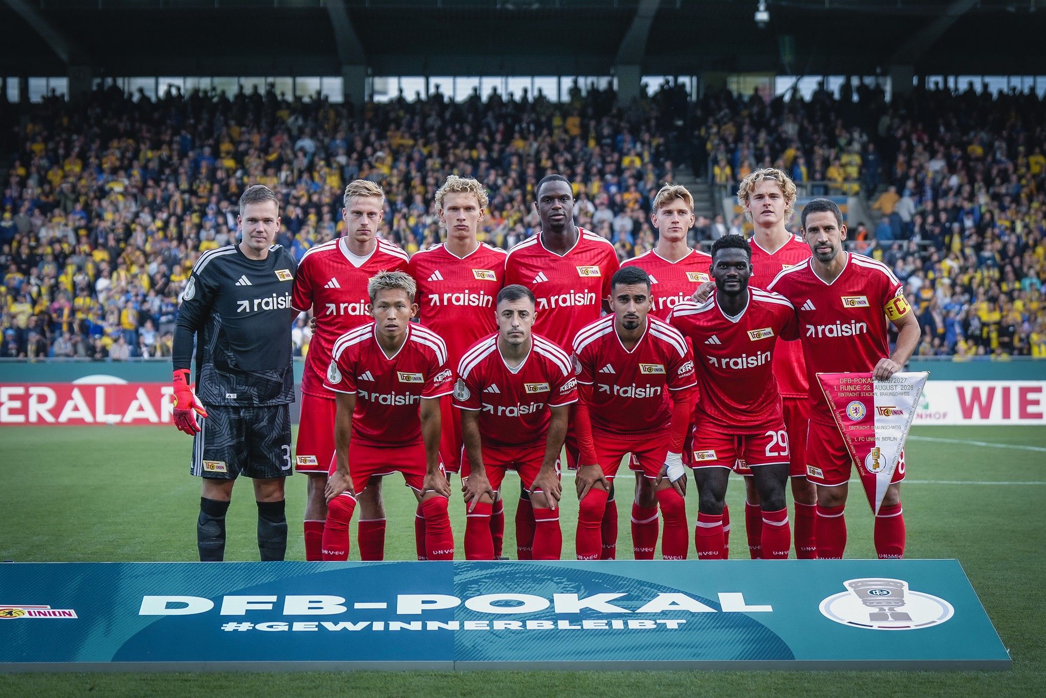 Eine Fußballmannschaft in roten Trikots steht auf dem Spielfeld für ein Gruppenfoto, mit einem Banner im Vordergrund.