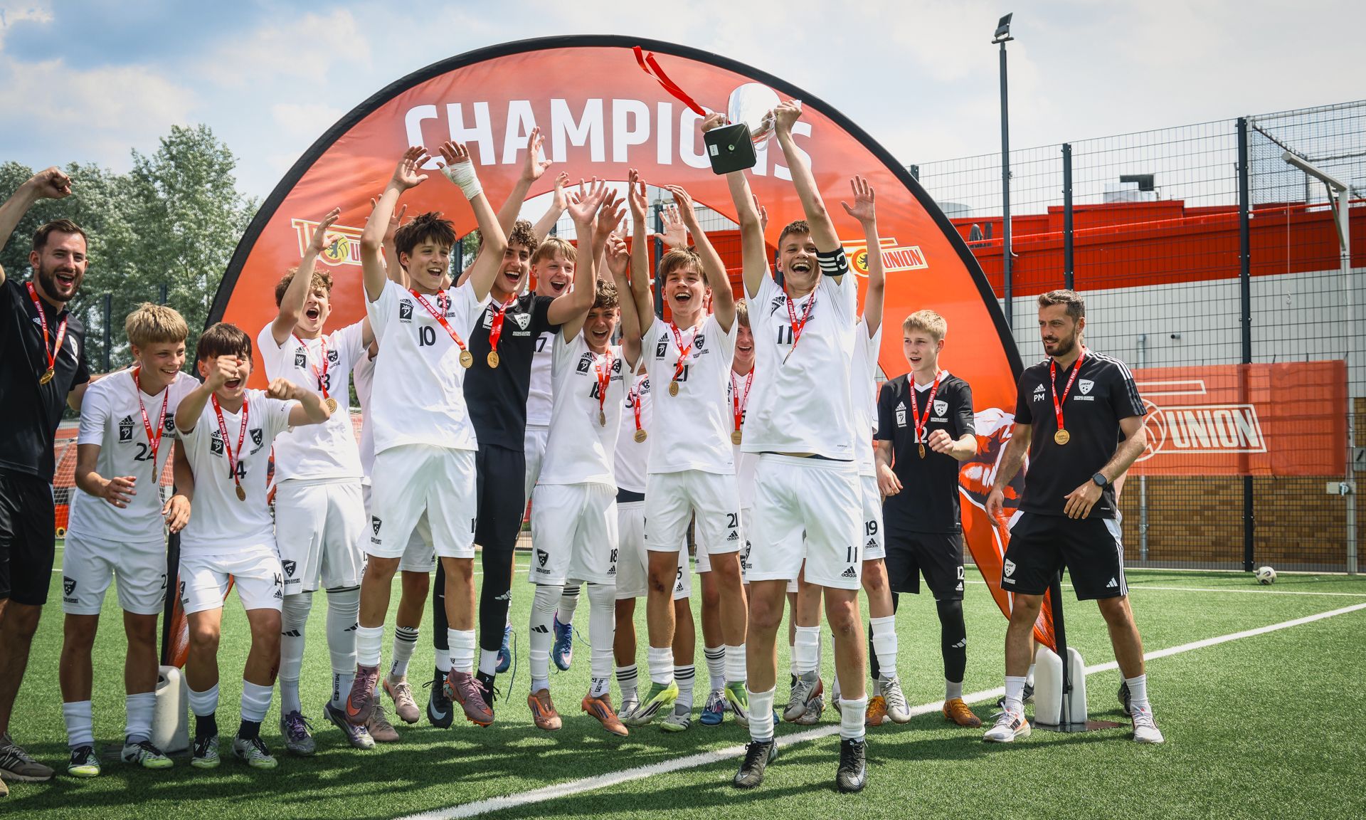 Jubelnde Fußballmannschaft mit Medaillen und Pokal vor einem roten Banner mit der Aufschrift "CHAMPIONS".