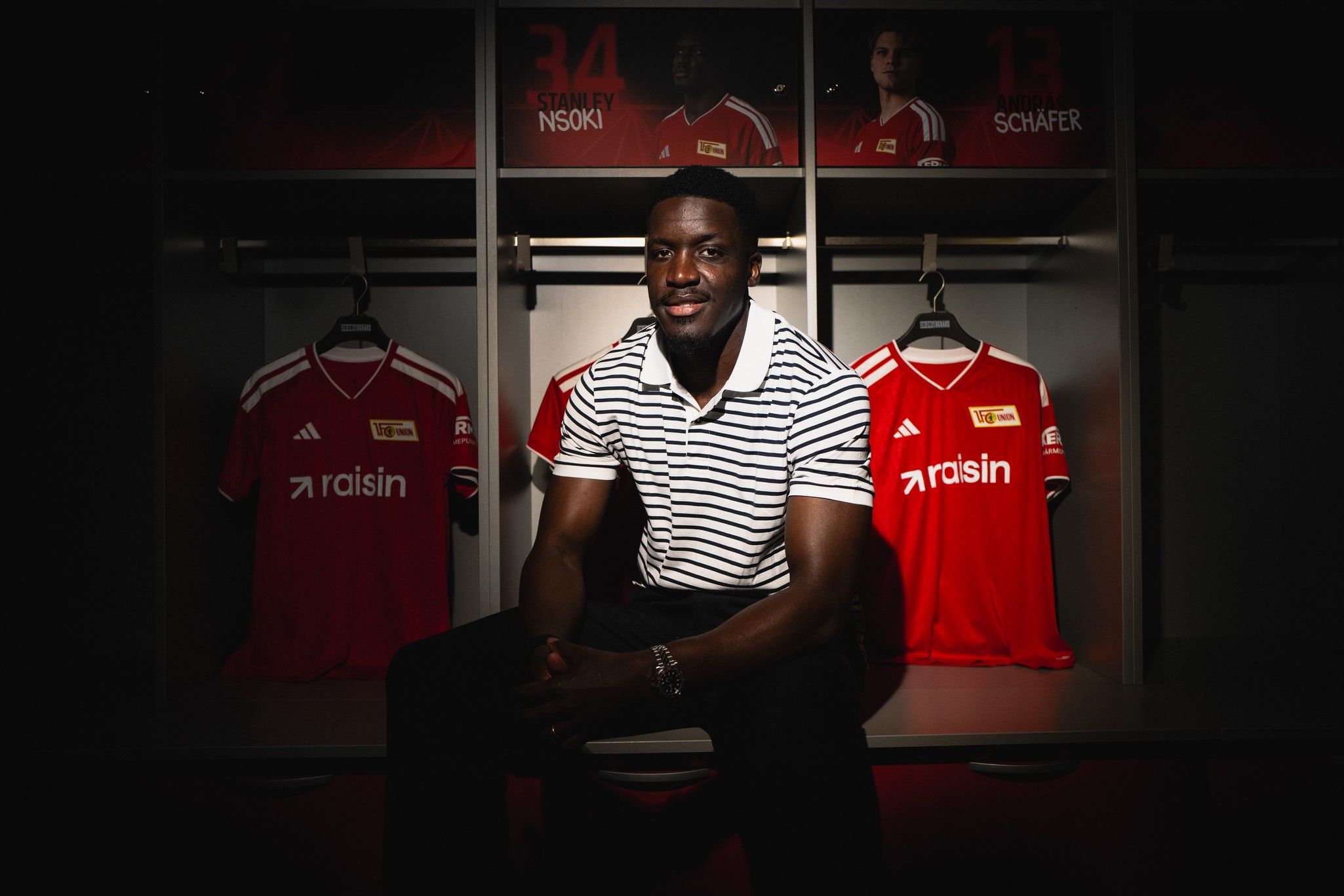 Ein Mann in einem gestreiften Shirt sitzt vor rot-weiß gestreiften Fußballtrikots in einem Umkleideraum.