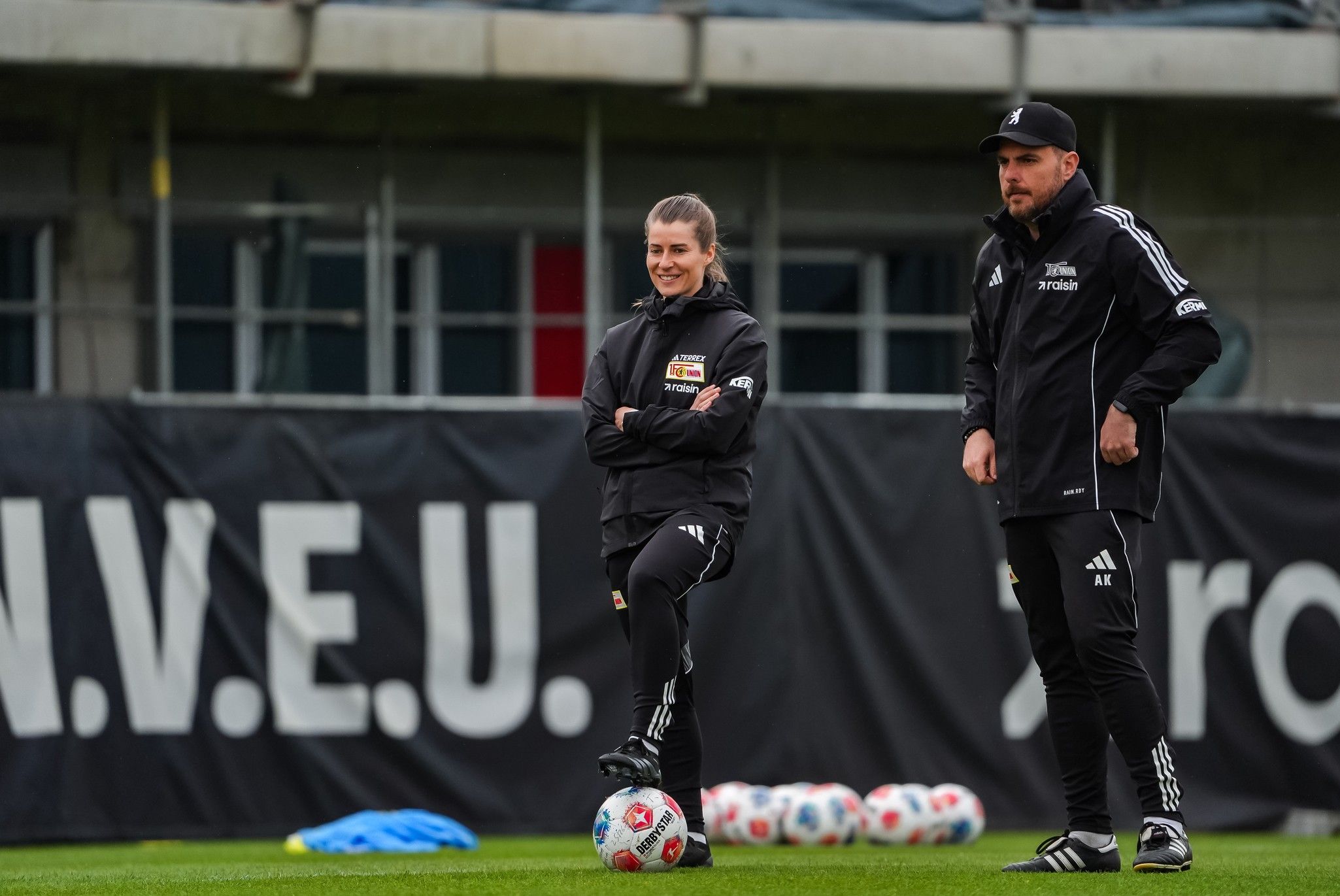 Eine Fußballspielerin steht mit einem Ball auf dem Trainingsplatz, daneben ein Trainer in schwarzer Sportkleidung.
