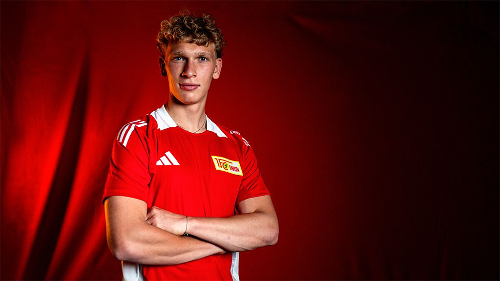 Young athlete in a red jersey with white accents stands with arms crossed in front of a red-lit background.