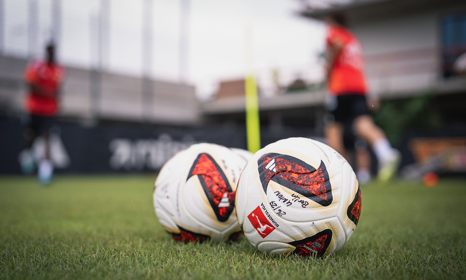 Zwei Fußballbälle liegen auf einem Trainingsplatz, im Hintergrund sind verschwommene Spieler in roten Trikots zu sehen.