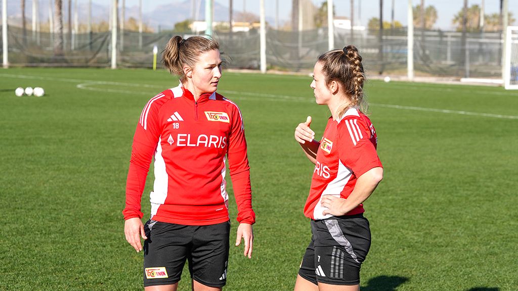 Zwei Fußballspielerinnen in roten Trainingsanzügen stehen auf einem grünen Platz und unterhalten sich.