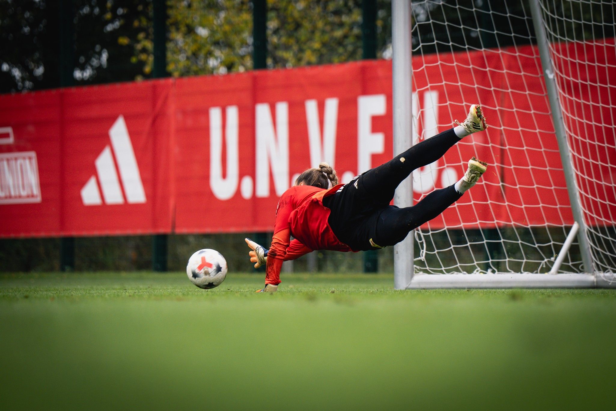 Torwart springt in die Luft, um einen Ball zu halten, während er vor einem Tor steht. Rotes Hintergrundbanner sichtbar.