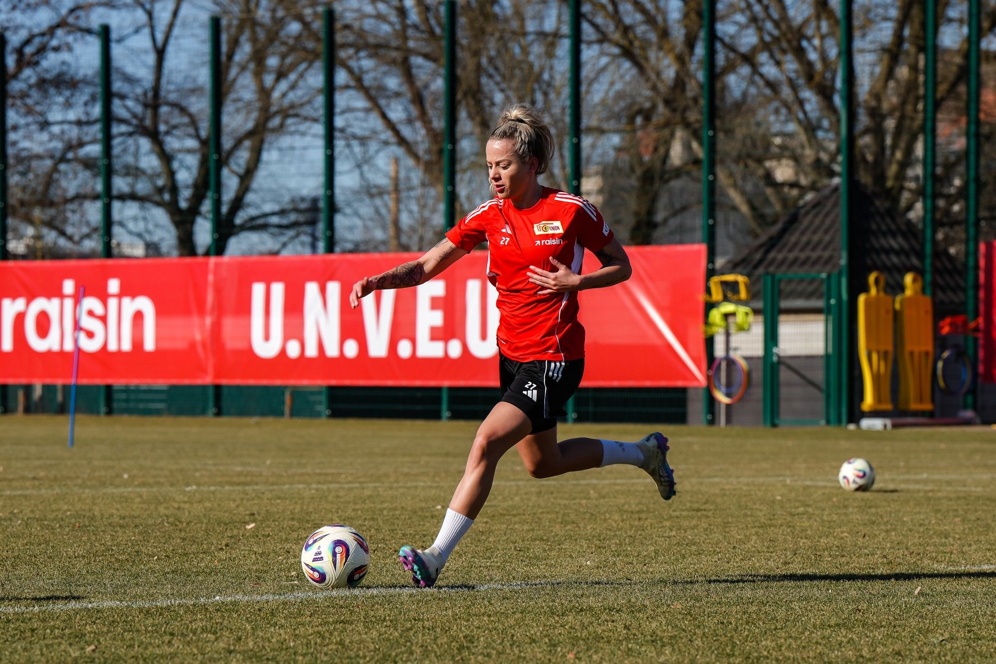 Eine Fußballspielerin dribbelt einen Ball auf einem Trainingsfeld, während sie in einem roten Trikot läuft. Im Hintergrund sind Trainingsgeräte sichtbar.