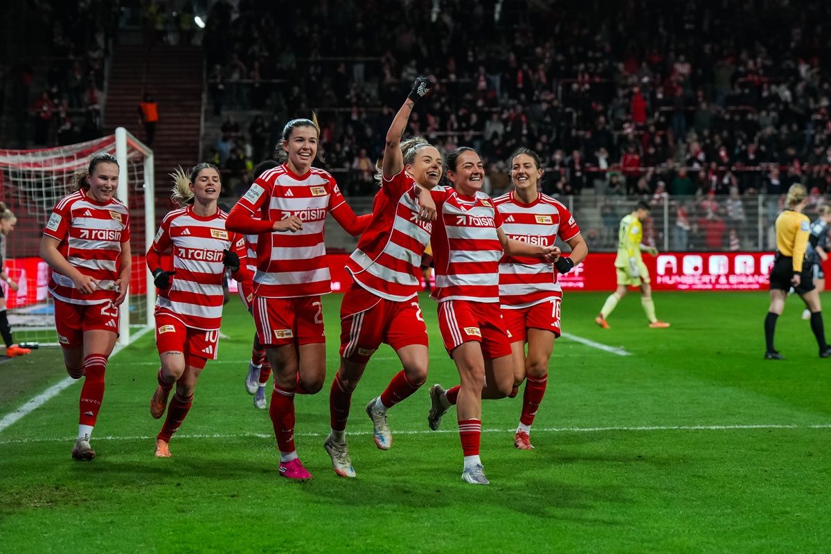 Frauenfußballmannschaft in roten Trikots feiert ein Tor, umgeben von jubelnden Teamkolleginnen auf dem Spielfeld.