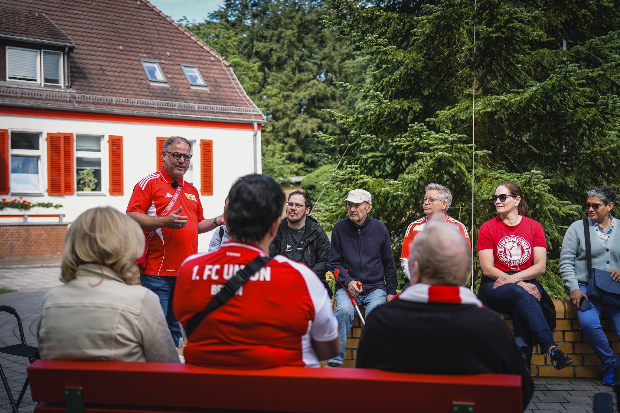 Eine Gruppe von sechs Personen sitzt im Freien, während ein Mann im roten Trikot spricht. Im Hintergrund sind Bäume und ein Gebäude sichtbar.