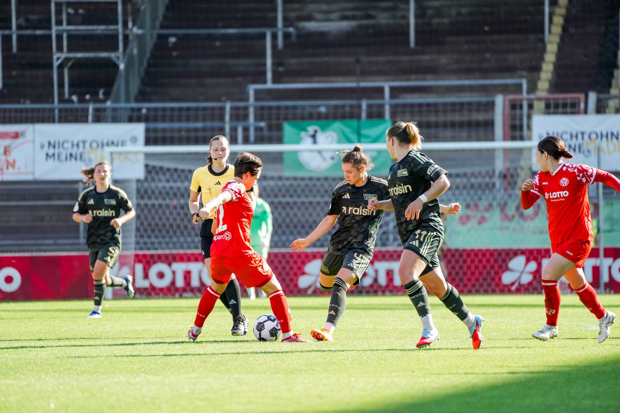 Zwei Frauenfußballspielerinnen im Wettkampf auf dem Spielfeld, mit einer weiteren Spielerin im Hintergrund und einem Torwart.