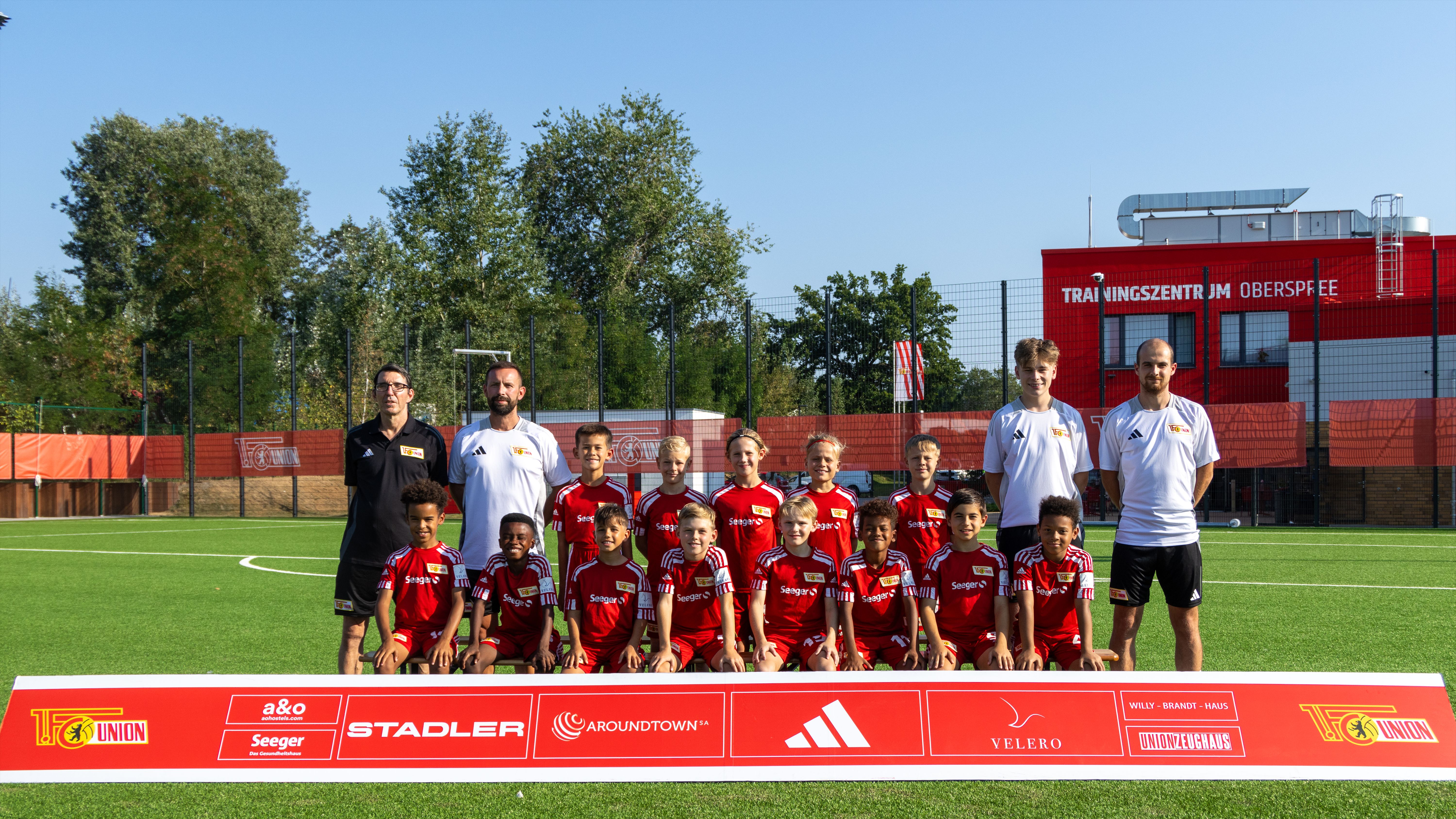 Mannschaftsbild einer Jugendfußballmannschaft auf einem Sportplatz, mit Trainern und Spielern in roten Trikots.