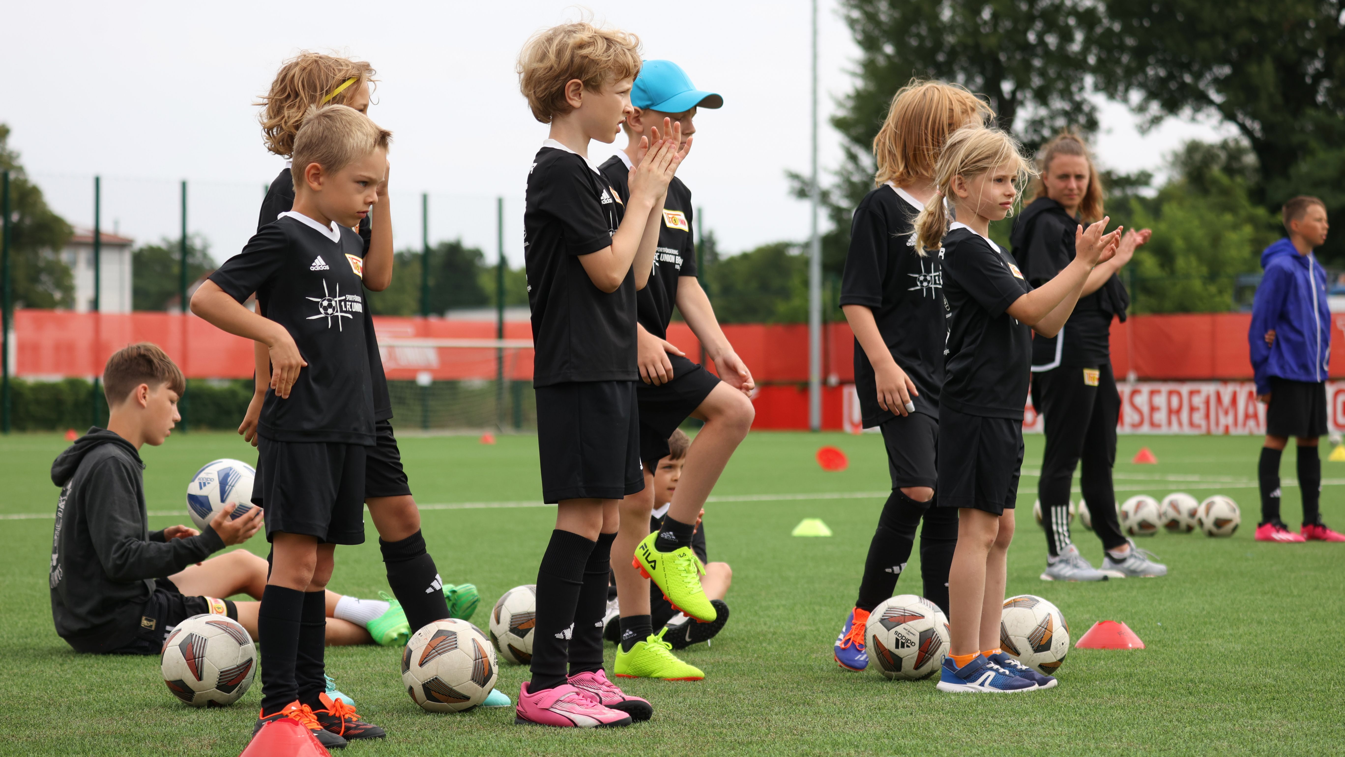 Eine Gruppe von Kindern in schwarzen Fußballtrikots steht auf einem Sportplatz, w�ährend sie einem Trainer zuhören.