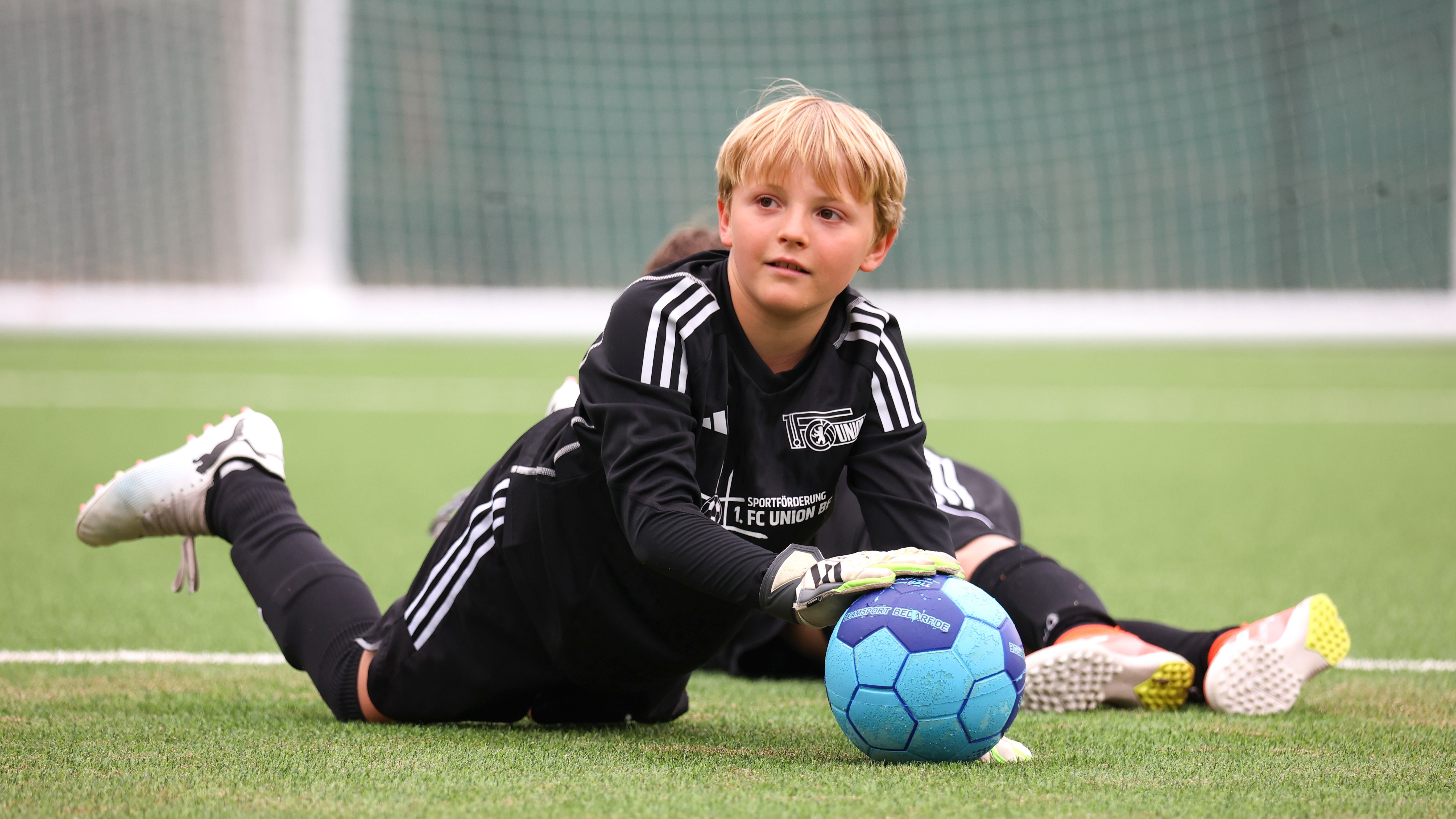 Ein Junge in schwarzer Fußballausrüstung liegt auf dem Rasen und hält einen blauen Ball, während ein weiterer Spieler im Hintergrund zu sehen ist.