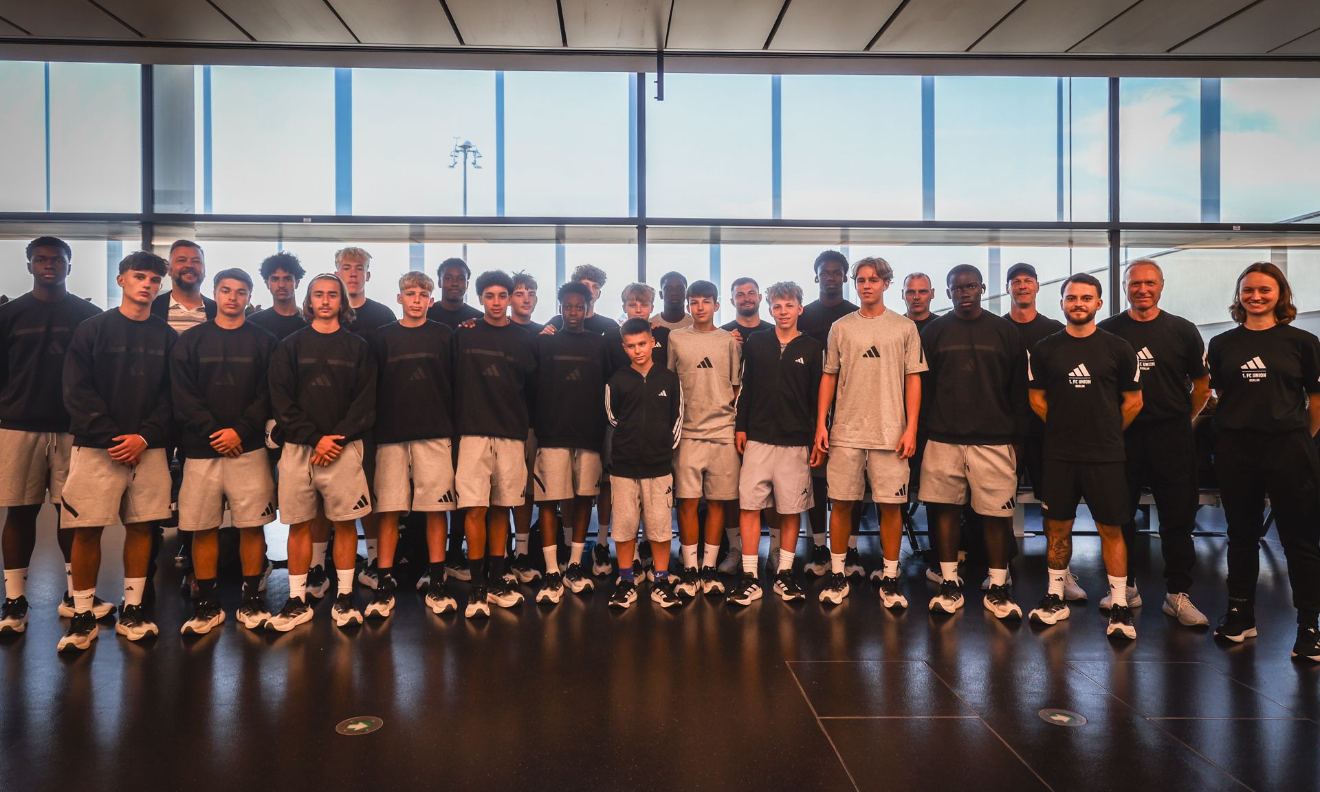 Gruppe von Basketballspielern in einheitlicher Sportkleidung steht vor großen Fenstern in einem modernen Raum.