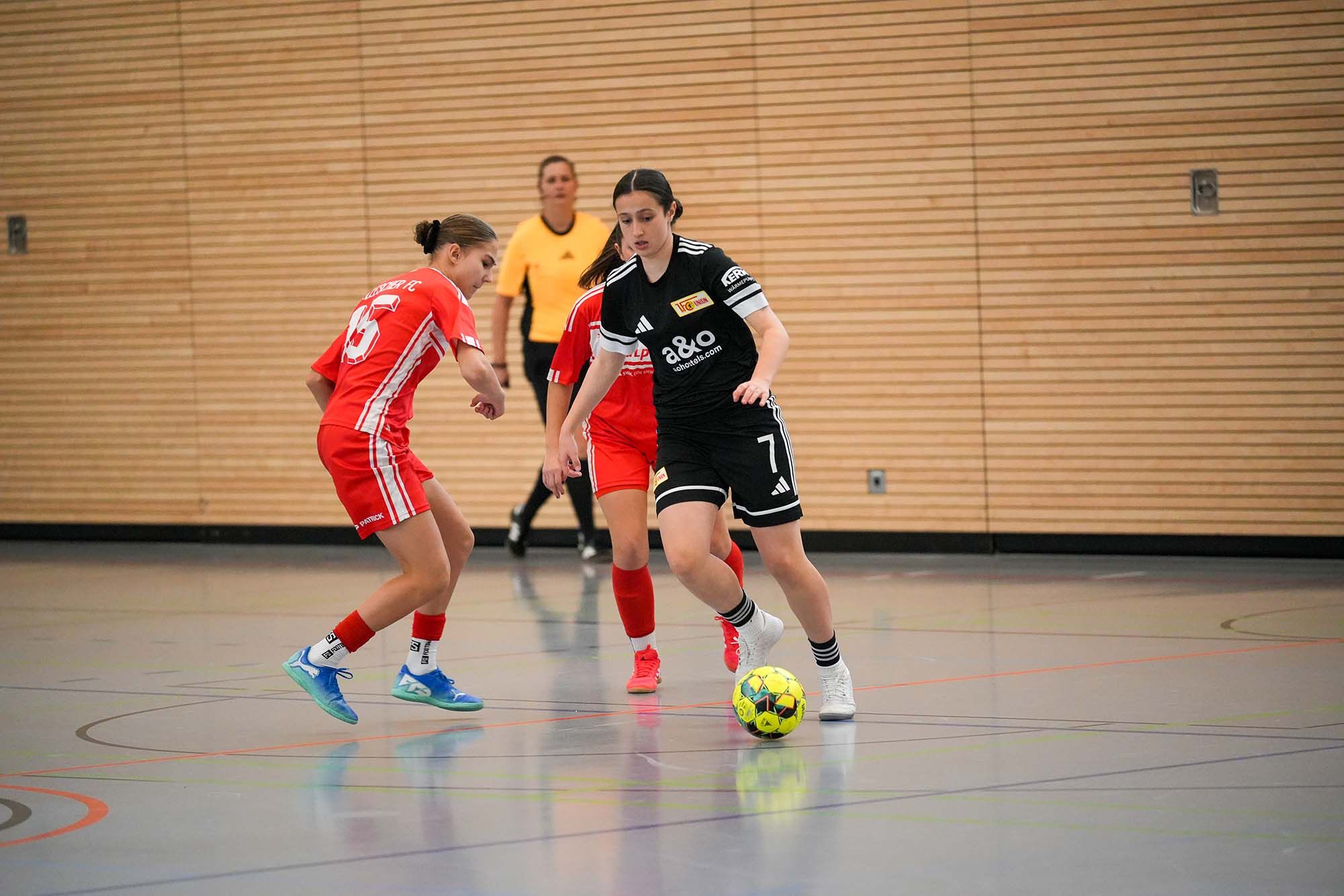 Zwei Frauen im Hallenfußball: eine in rotem Trikot dribbelt, die andere in schwarzem Trikot mit der Nummer 7 verteidigt.