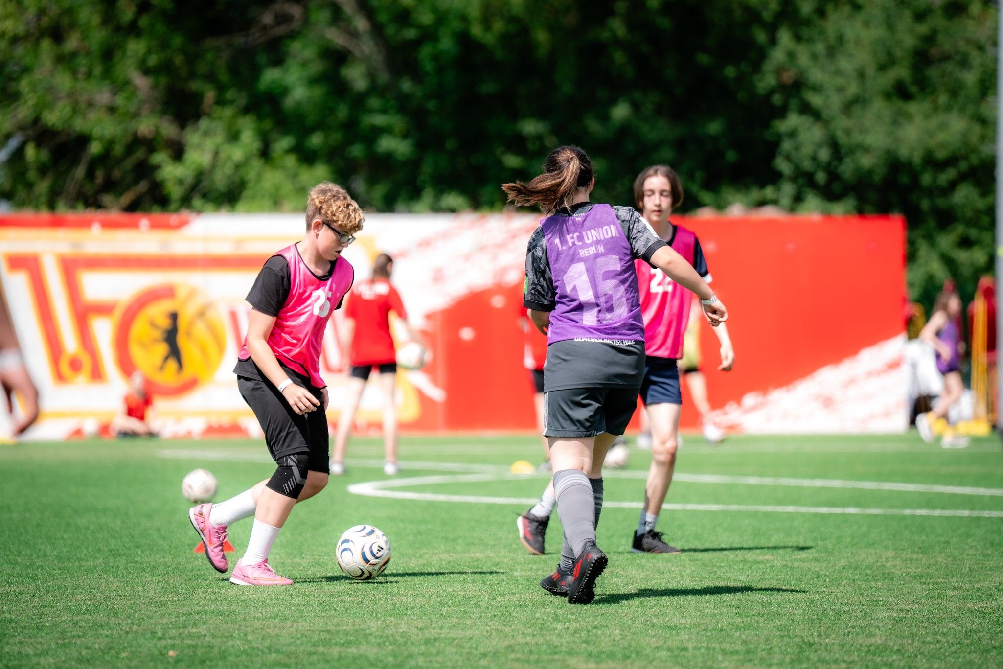 Zwei Spieler in pinken Trikots dribbeln auf einem Fußballfeld, während ein Torwart im Hintergrund bereitsteht.