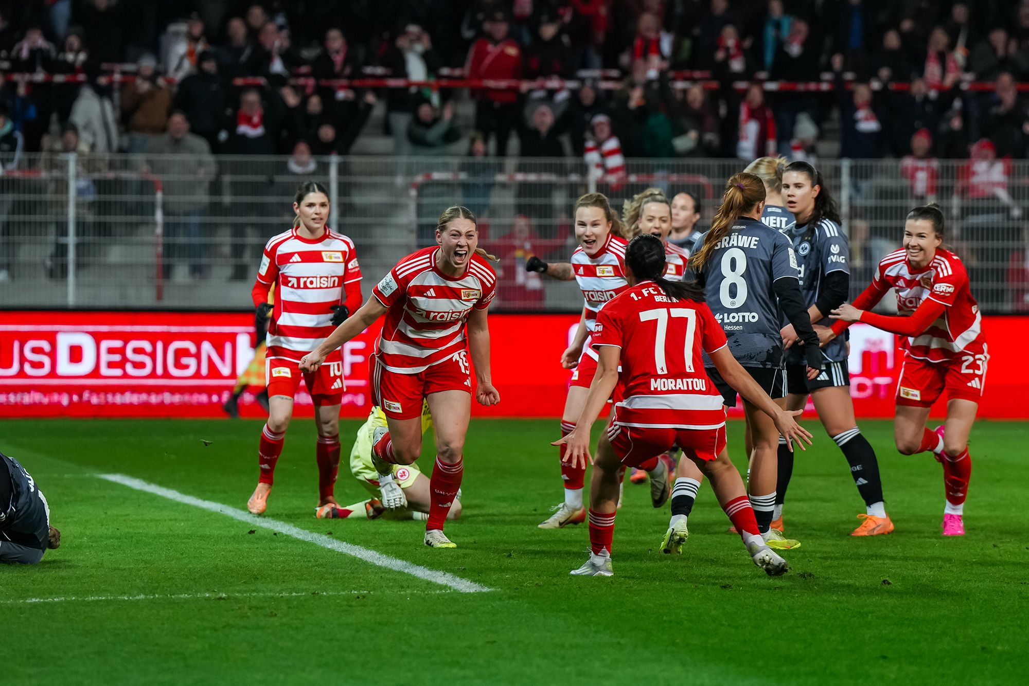 Feiernde Fußballspielerinnen in roten Trikots jubeln nach einem Tor auf dem Spielfeld, umgeben von Fans im Stadion.