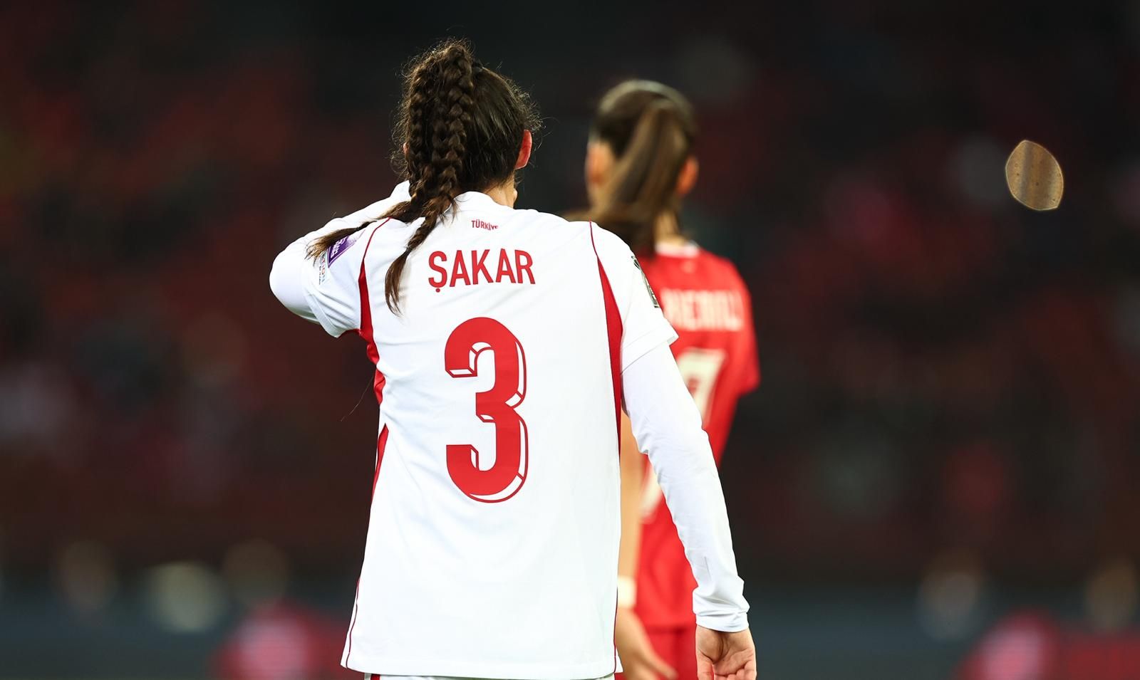 Eine Spielerin in einem weißen Fußballtrikot mit der Nummer 3 und dem Namen "Şakar" steht im Vordergrund, im Hintergrund eine weitere Spielerin.