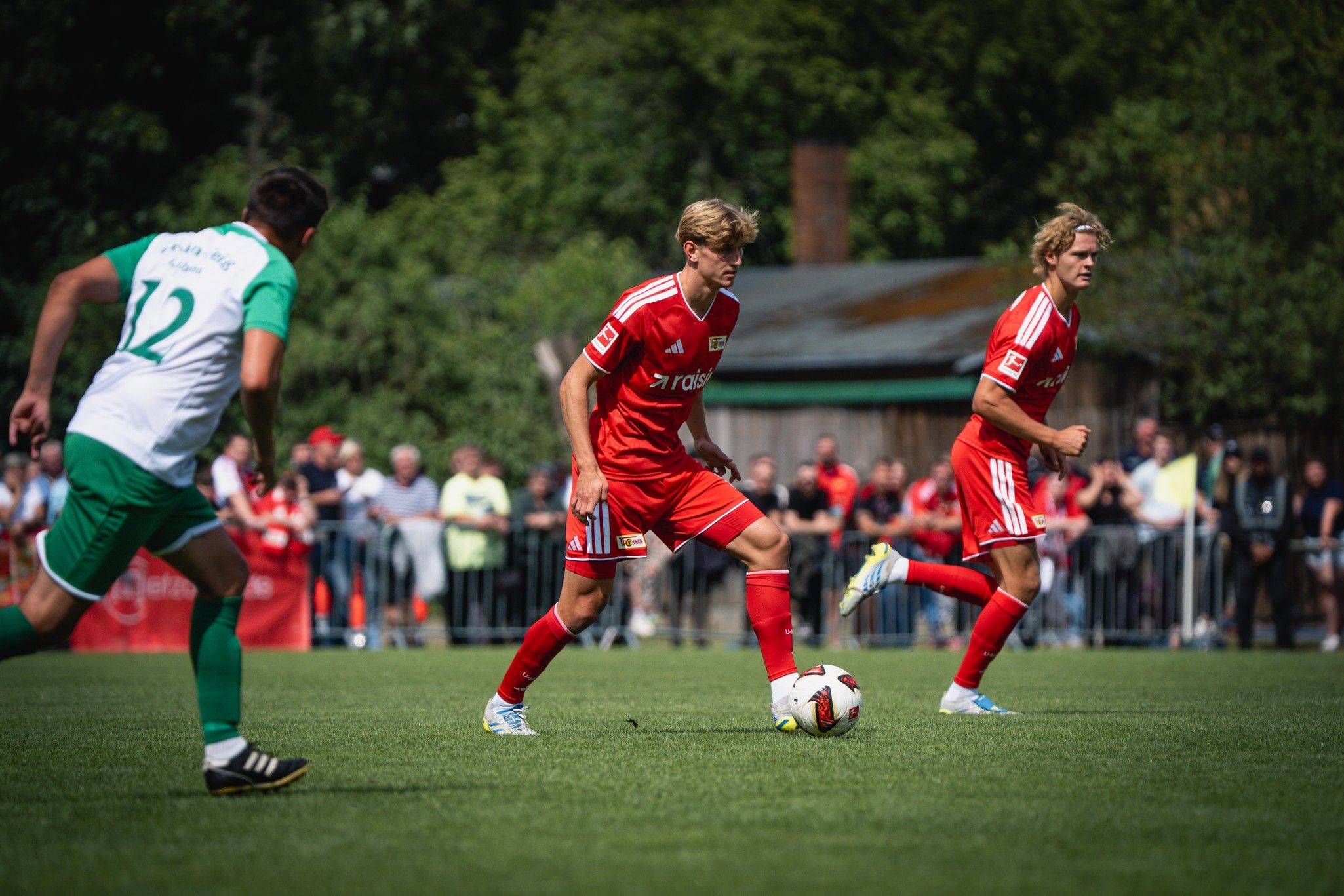 Zwei Fußballspieler in roten Trikots dribbeln den Ball auf einem Spielfeld, während ein Verteidiger in grünem Trikot hinter ihnen steht.