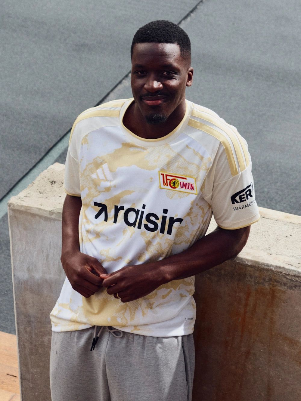 Ein lächelnder Mann in einem Fußballtrikot mit dem Logo von 1. FC Union und dem Schriftzug "raisin".