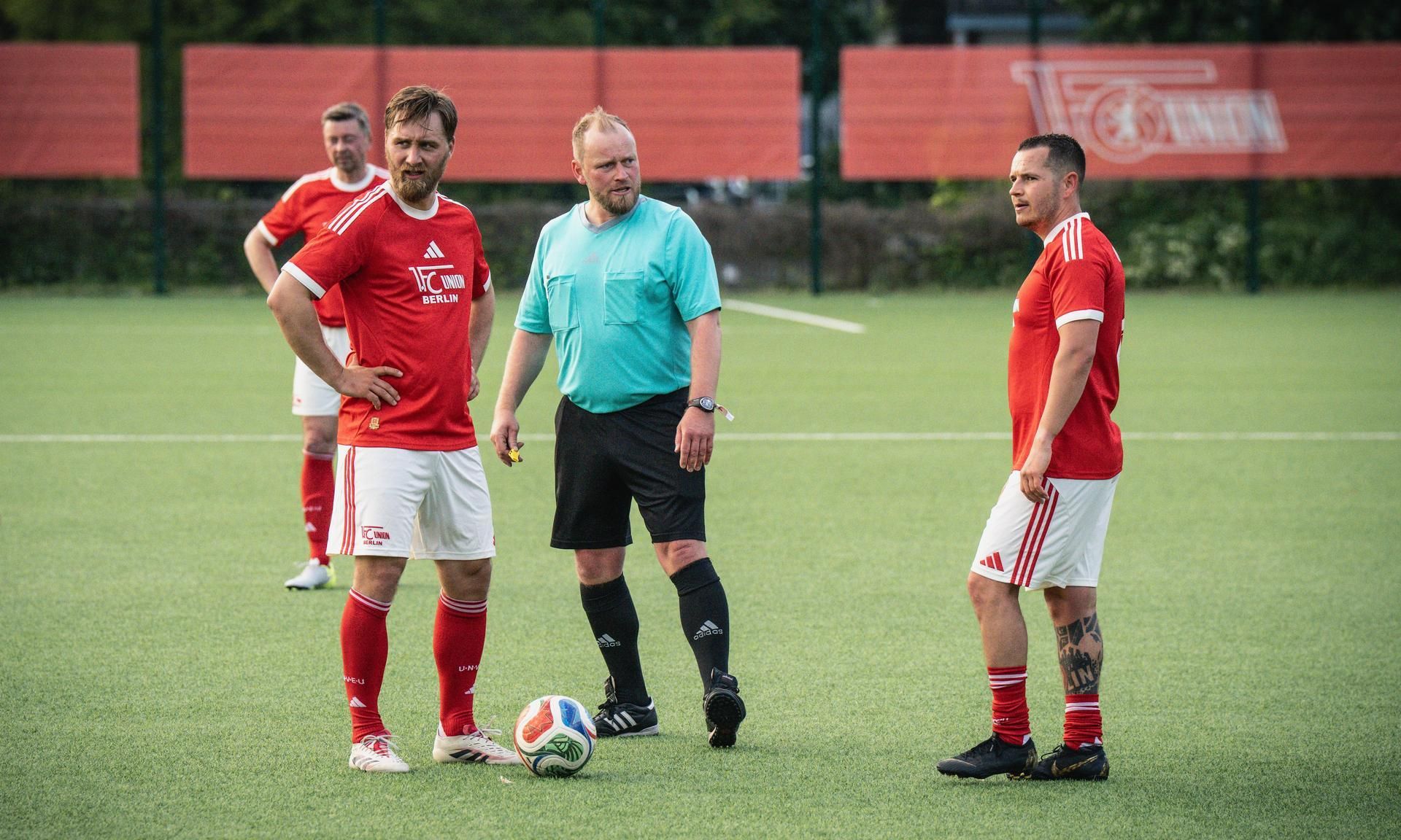Drei Fußballspieler in roten Trikots stehen auf einem Kunstrasenplatz, während ein Schiedsrichter zwischen ihnen steht.