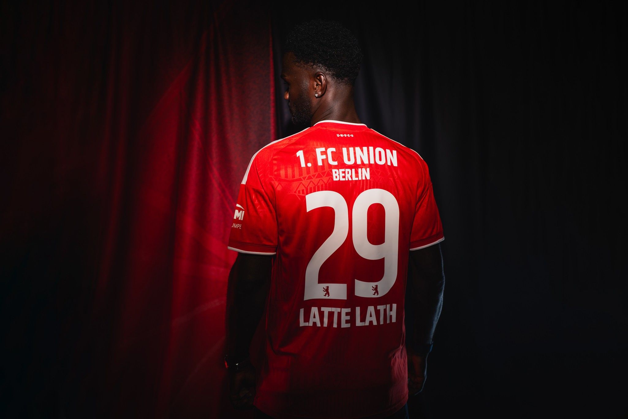 Ein Mann mit Rückenansicht trägt ein rotes Fußballtrikot des 1. FC Union Berlin mit der Nummer 29 und dem Namen "Latte Lath".